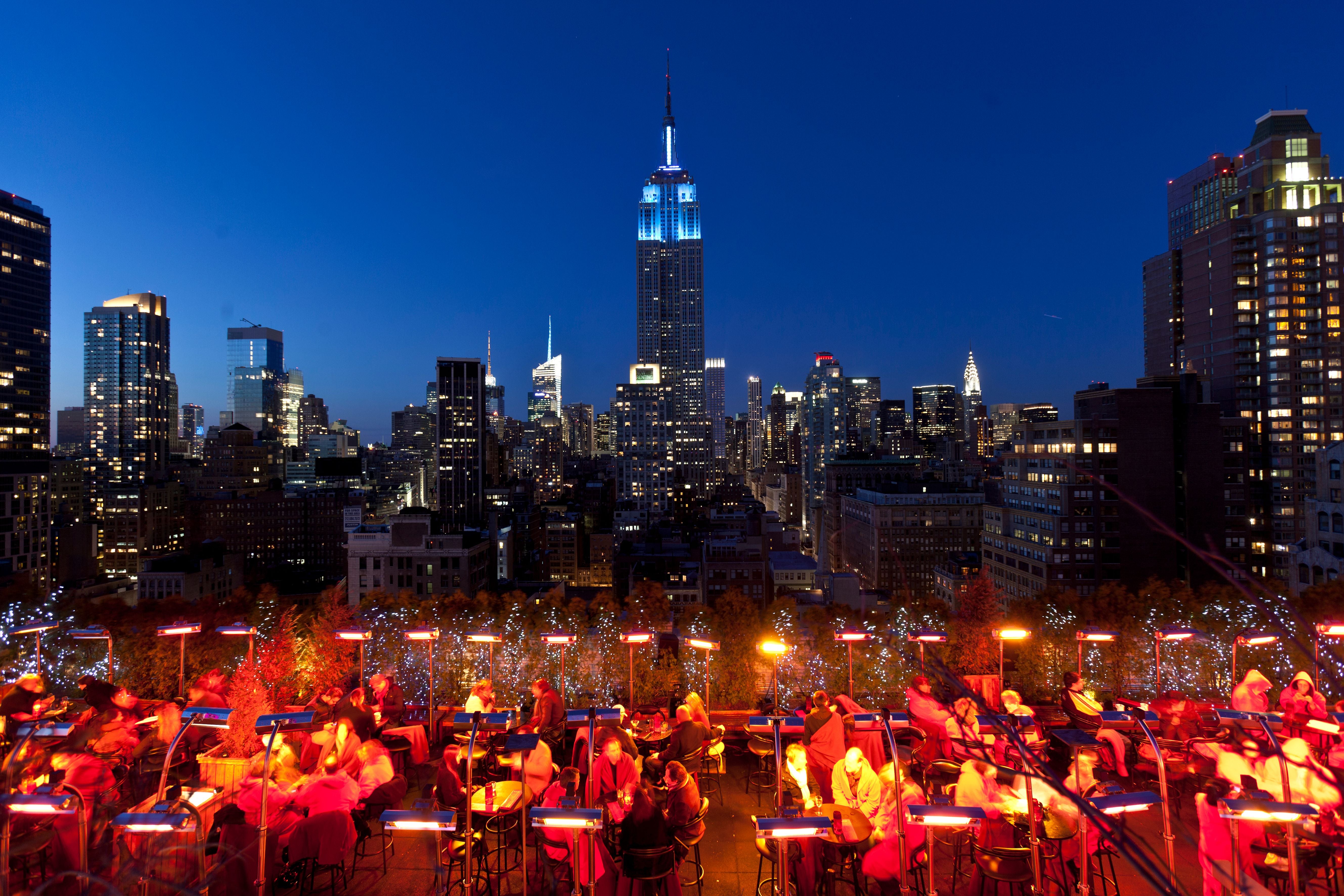 Menschen sitzen abends auf einer Dachterrasse mit Blick auf das Empire State Building in Manhattan.