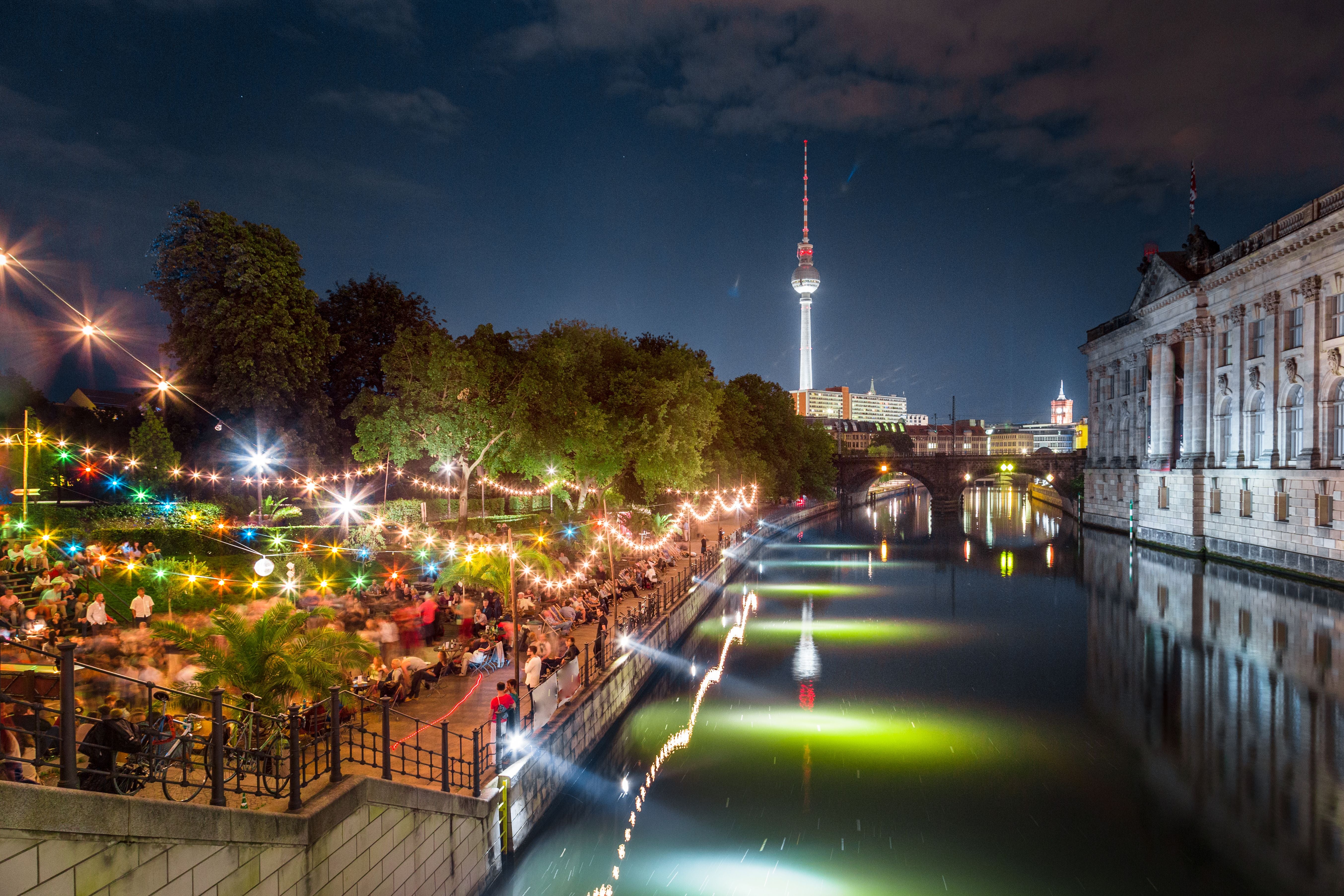 Menschen feiern abends an der Strandbar an der Spree mit Fernsehturm und Museumsinsel in Berlin.