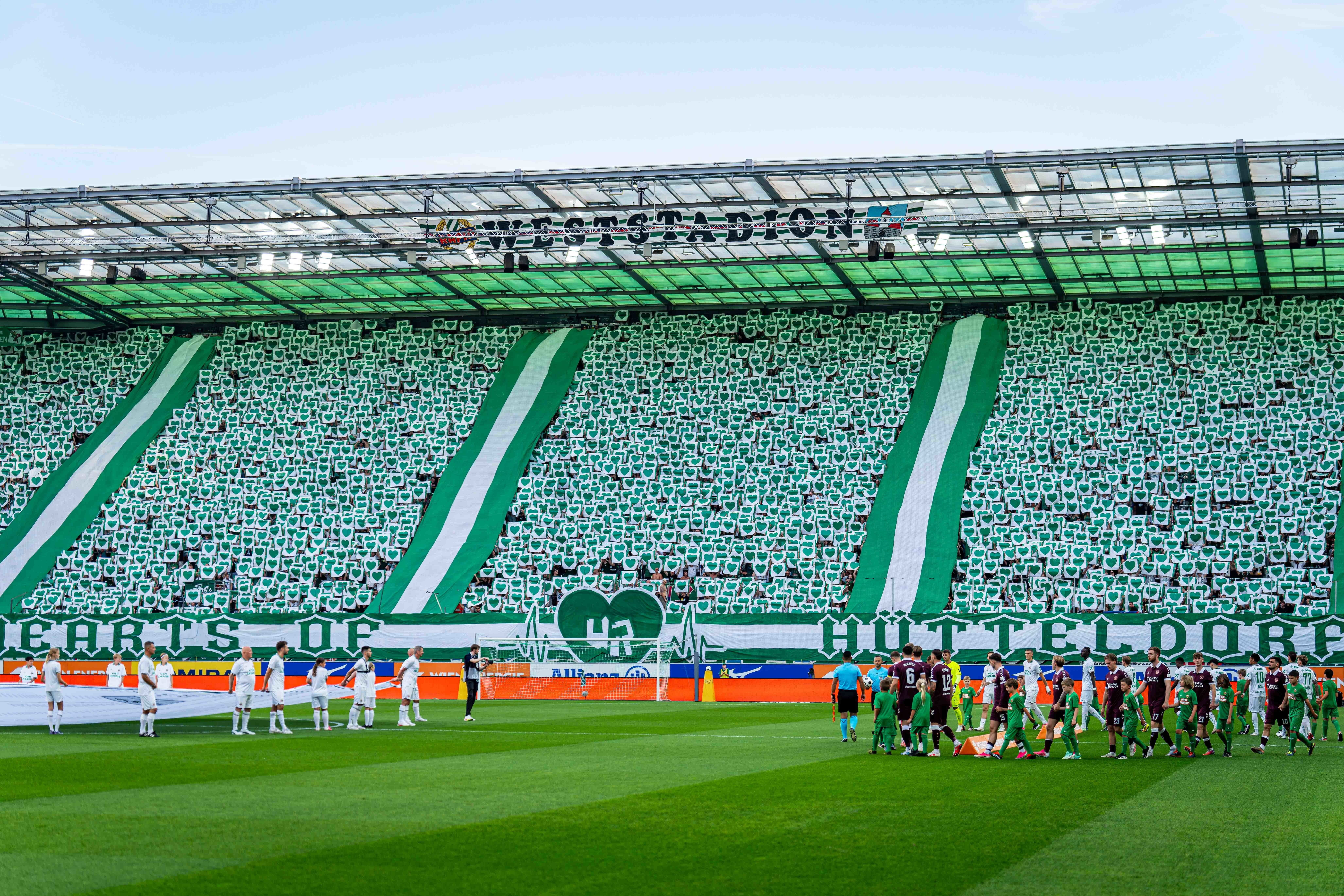 Fußballspieler betreten das Spielfeld vor einer grünen Fankurve mit Choreografie im Stadion.