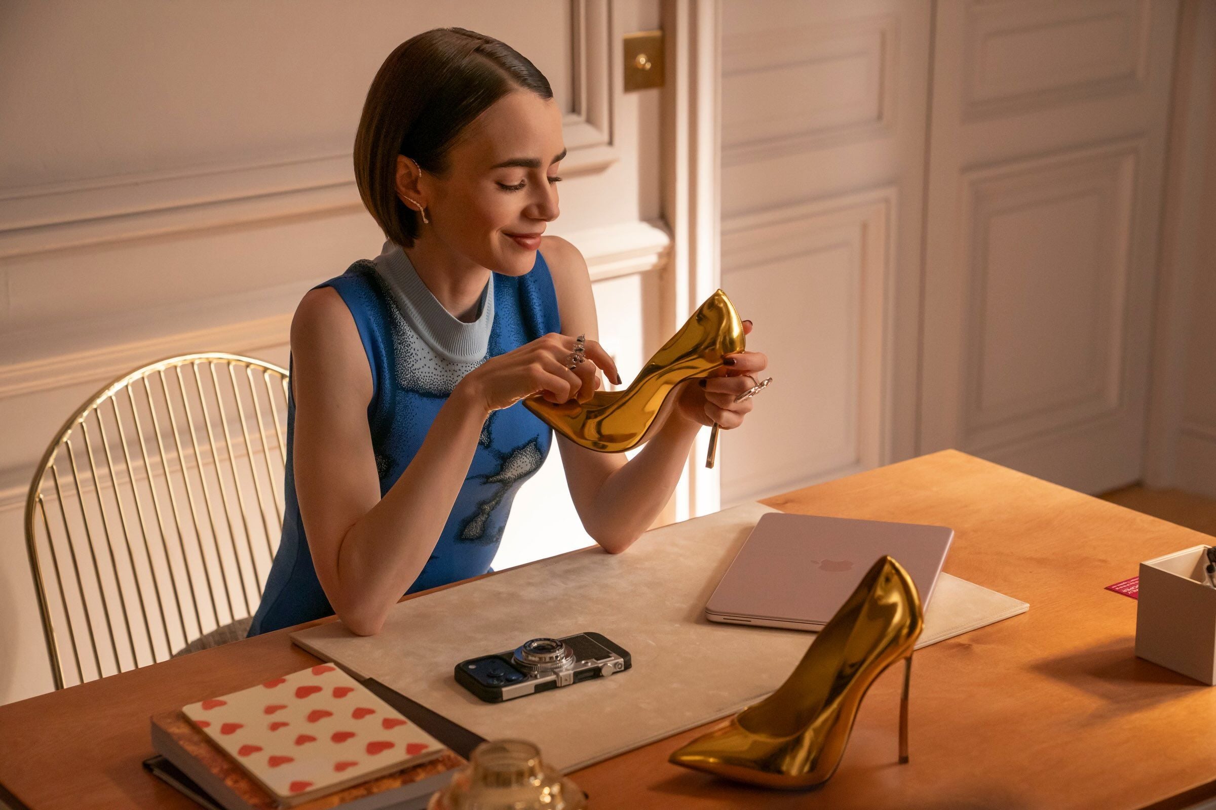 Frau sitzt am Tisch und betrachtet einen goldenen High Heel, zweites Paar steht daneben.