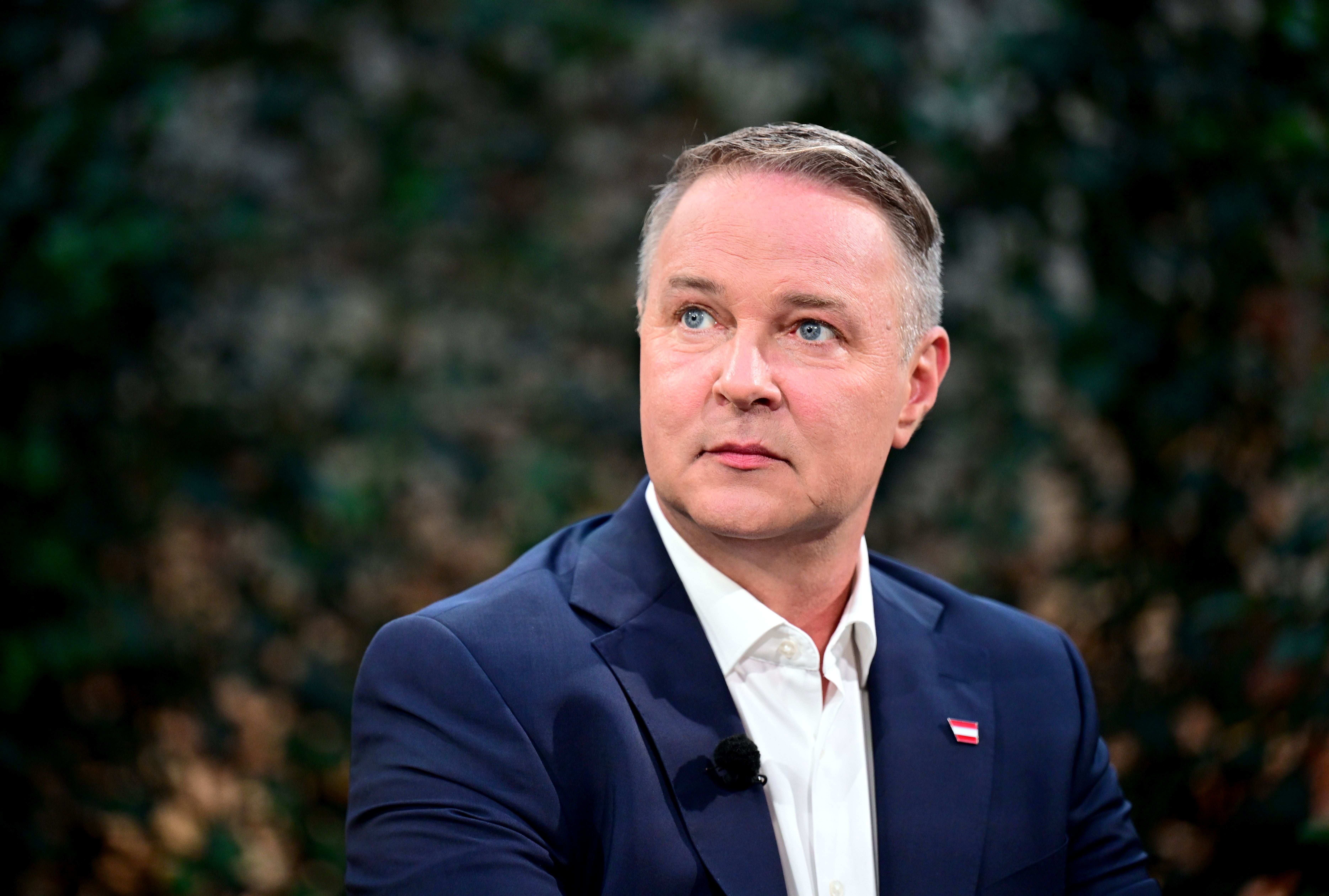 Ein Mann im blauen Anzug mit weißem Hemd sitzt vor unscharfem Hintergrund und blickt zur Seite.