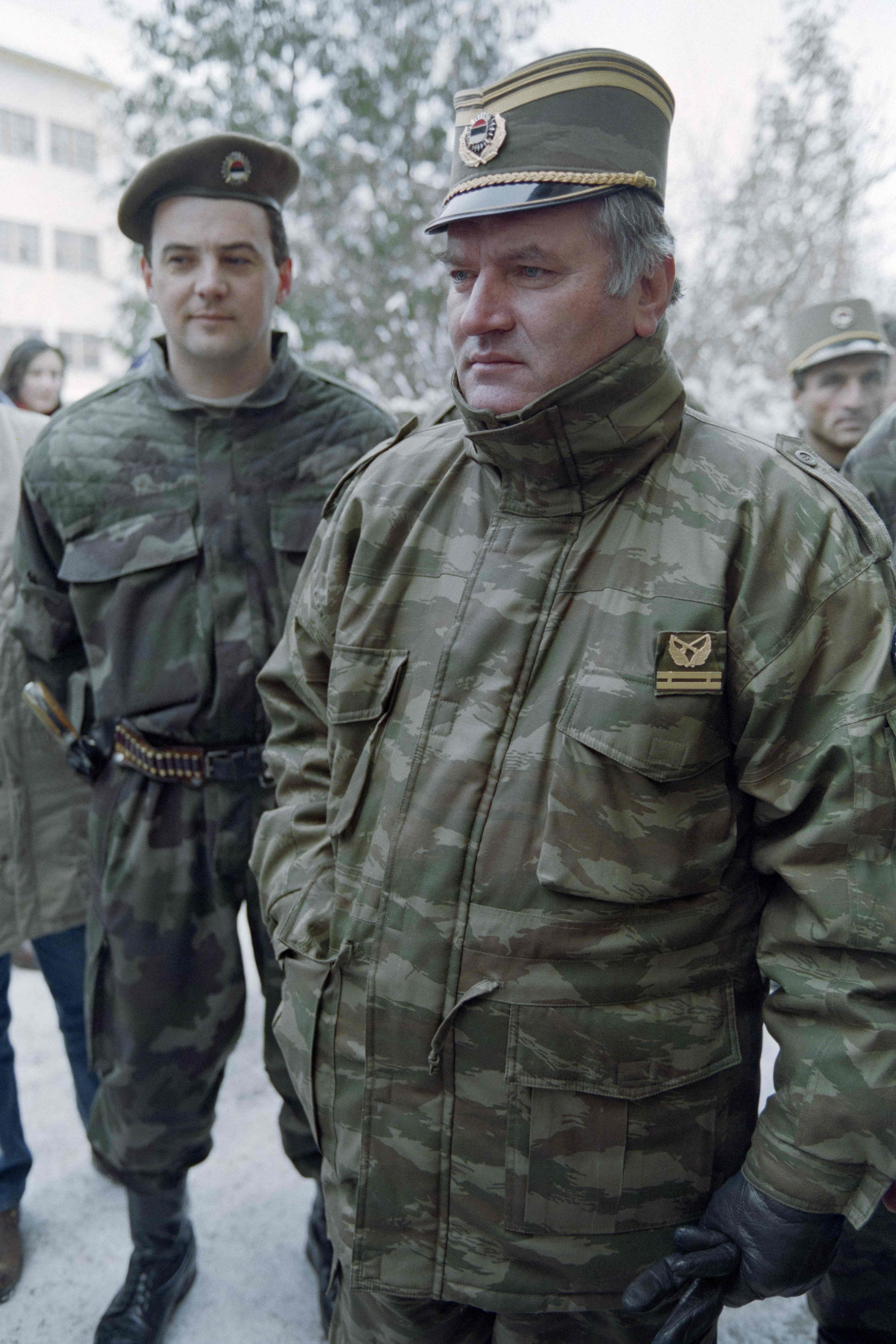 Zwei Männer in Militäruniformen stehen im Freien, einer trägt schwarze Handschuhe und eine Schirmmütze.