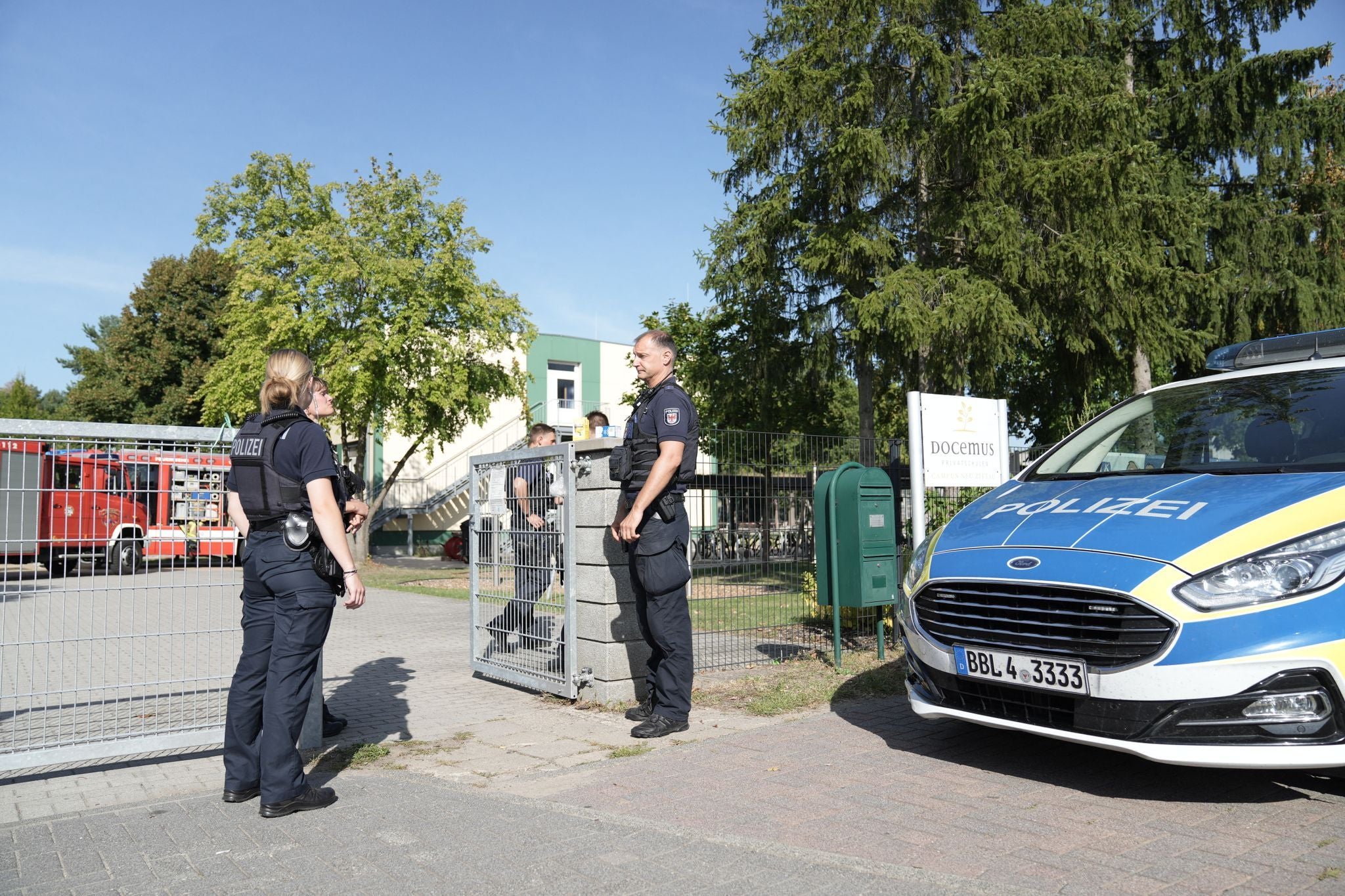 Zwei Polizisten stehen vor einem Schultor neben einem Polizei- und Feuerwehrfahrzeug.