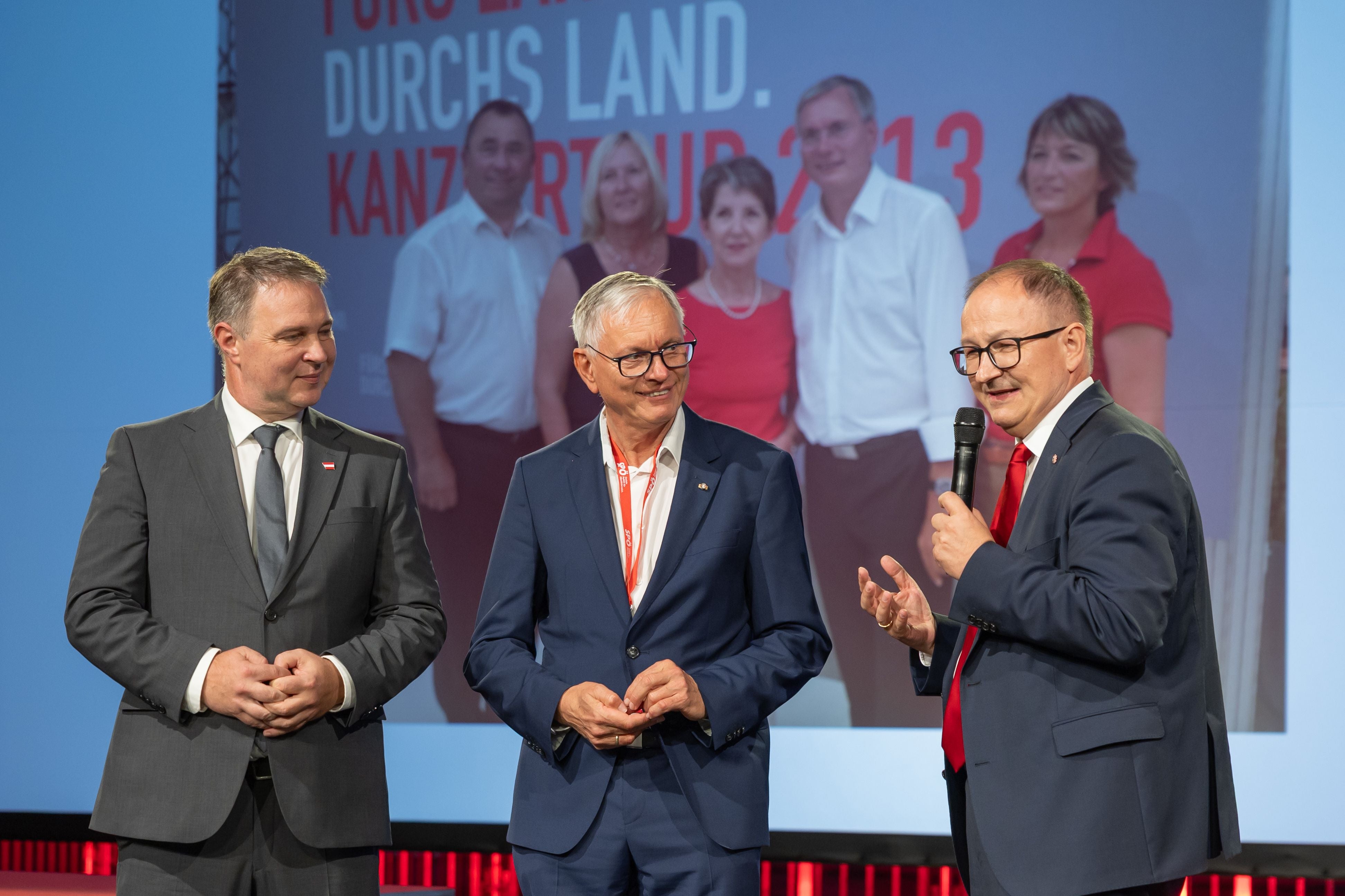 Drei Männer im Anzug sprechen auf einer Bühne, im Hintergrund ein Großbild mit weiteren Personen.