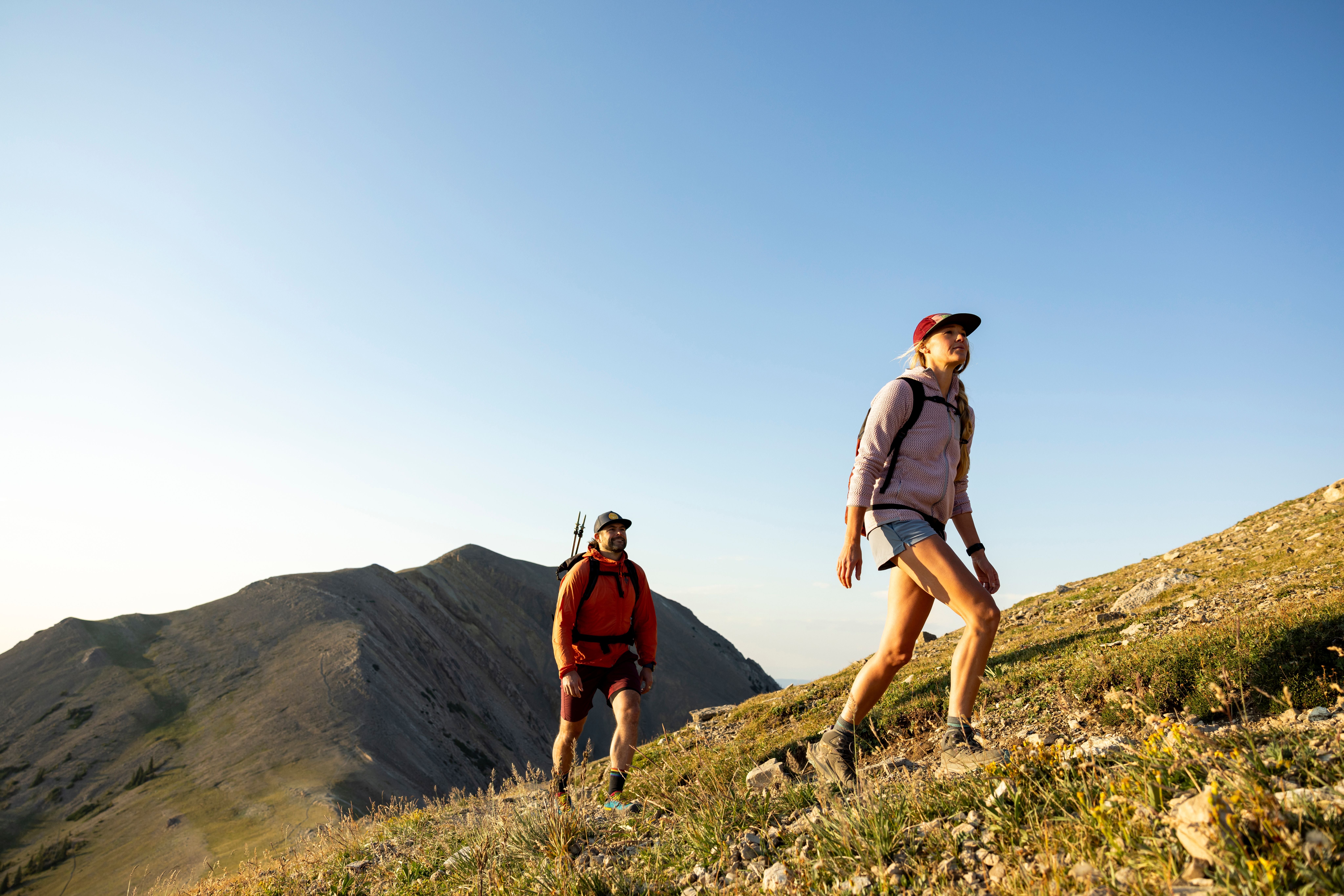 Zwei Menschen wandern bei sonnigem Wetter einen grasbewachsenen Berghang hinauf.