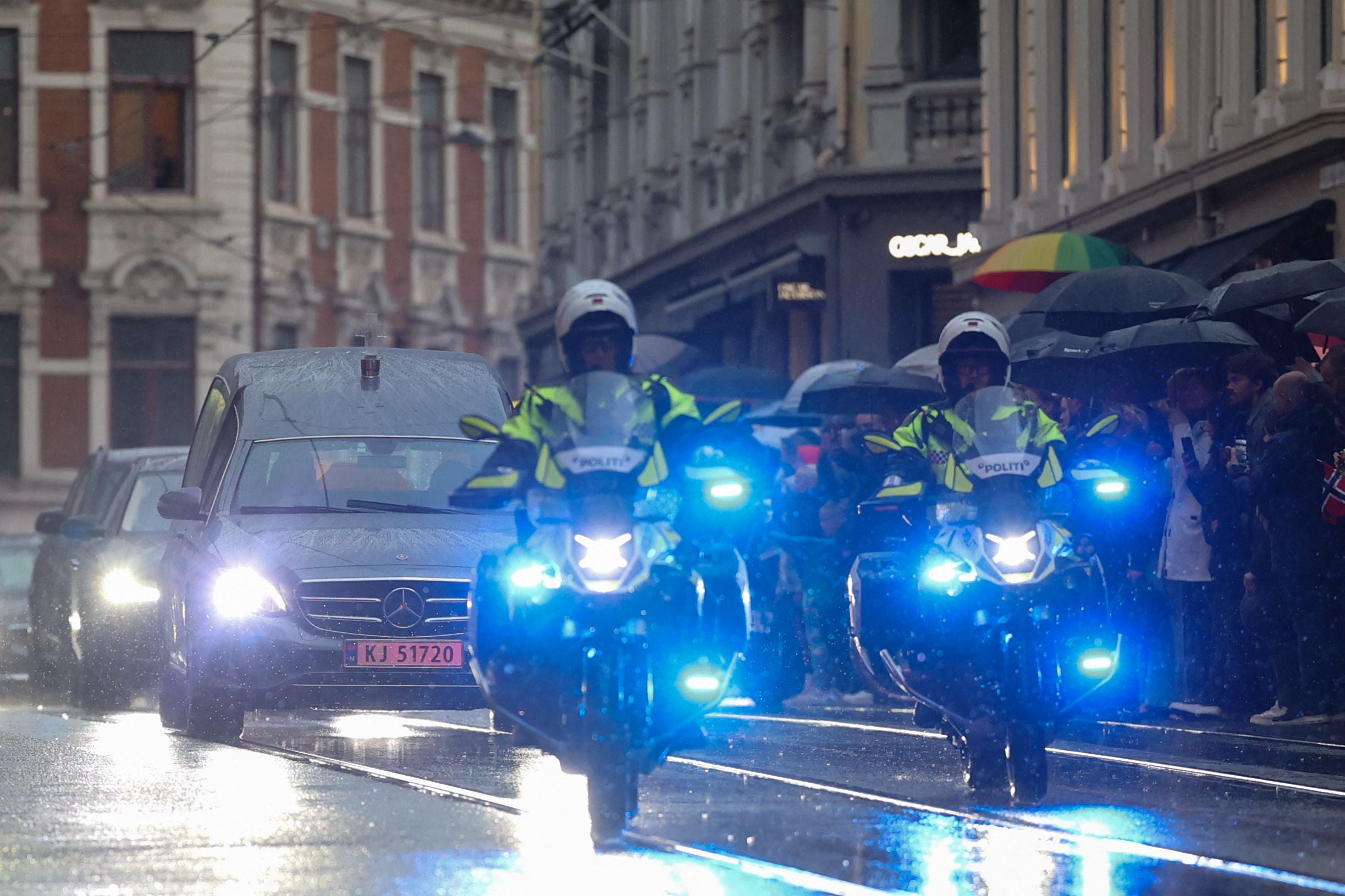 Polizeieskorte begleitet einen Leichenwagen in Oslo, während Menschen im Regen am Straßenrand stehen.