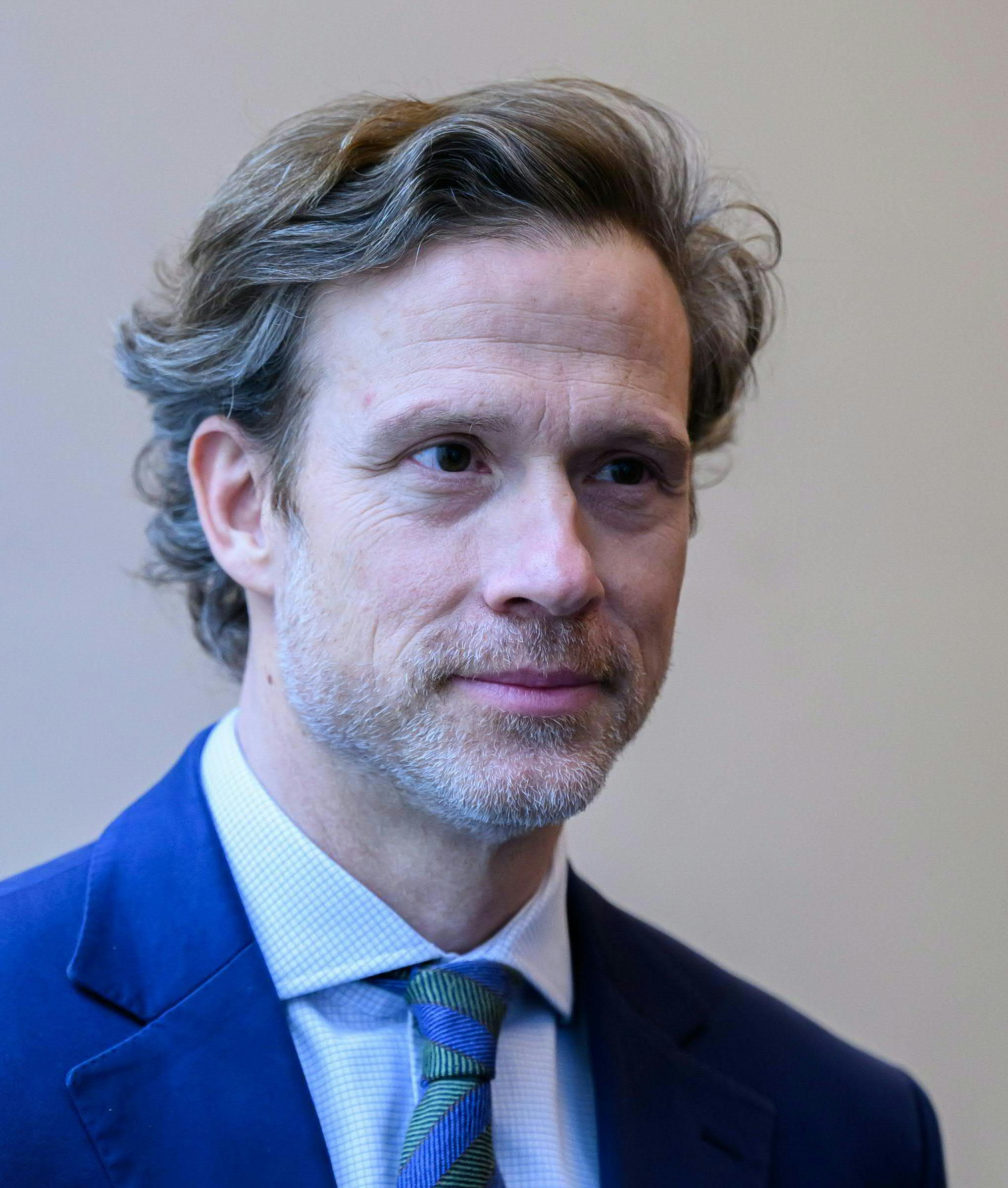 Ein Mann im blauen Anzug und gestreifter Krawatte blickt leicht lächelnd nach vorn.