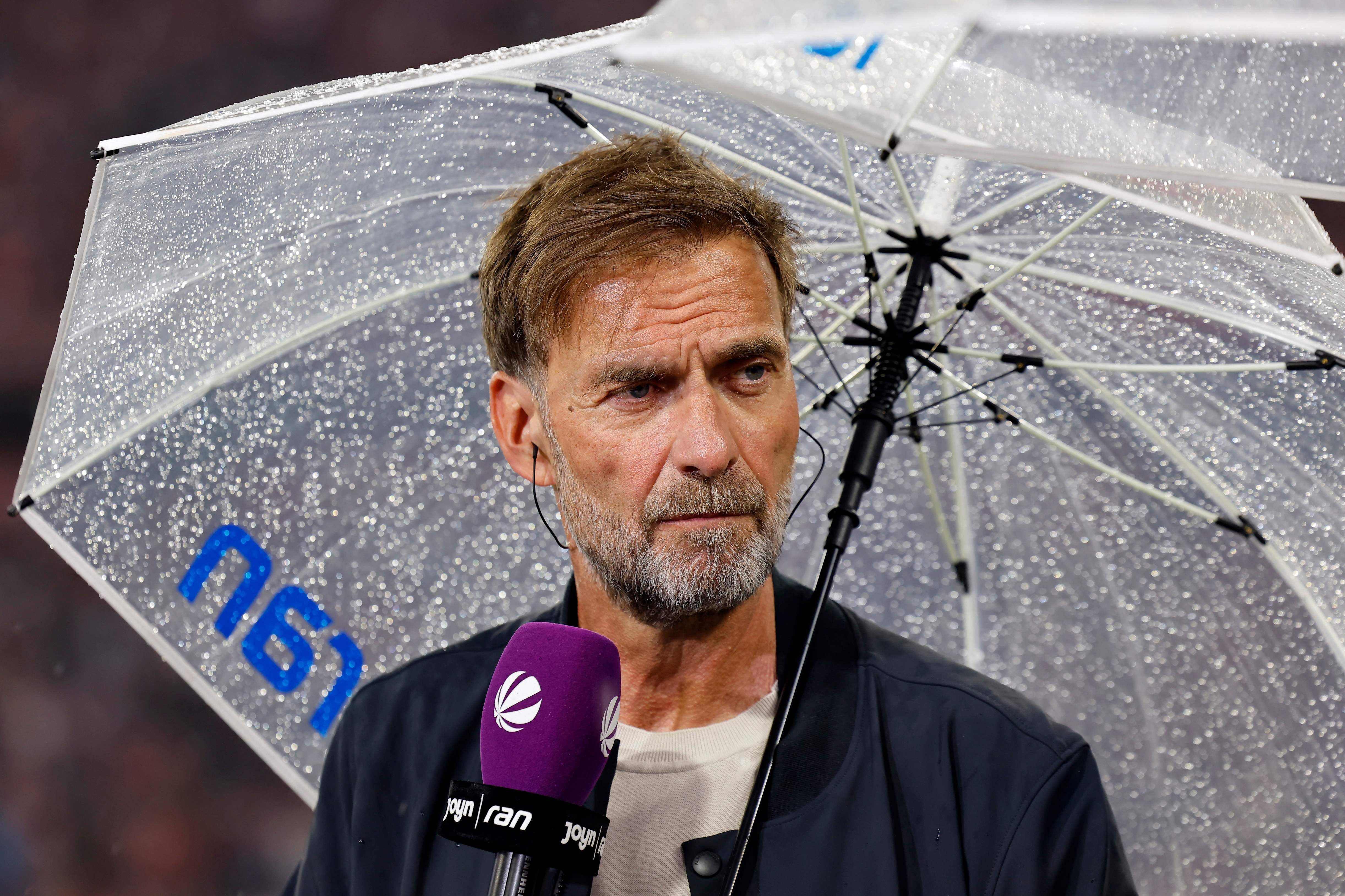 Ein Mann mit Bart hält einen Regenschirm und spricht in ein Mikrofon bei Regenwetter.