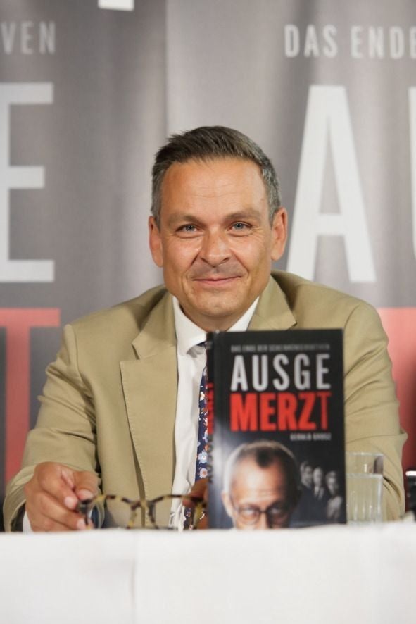 Ein Mann im Anzug sitzt hinter einem Buch mit dem Titel "AUSGEMERZT" auf einem Tisch.