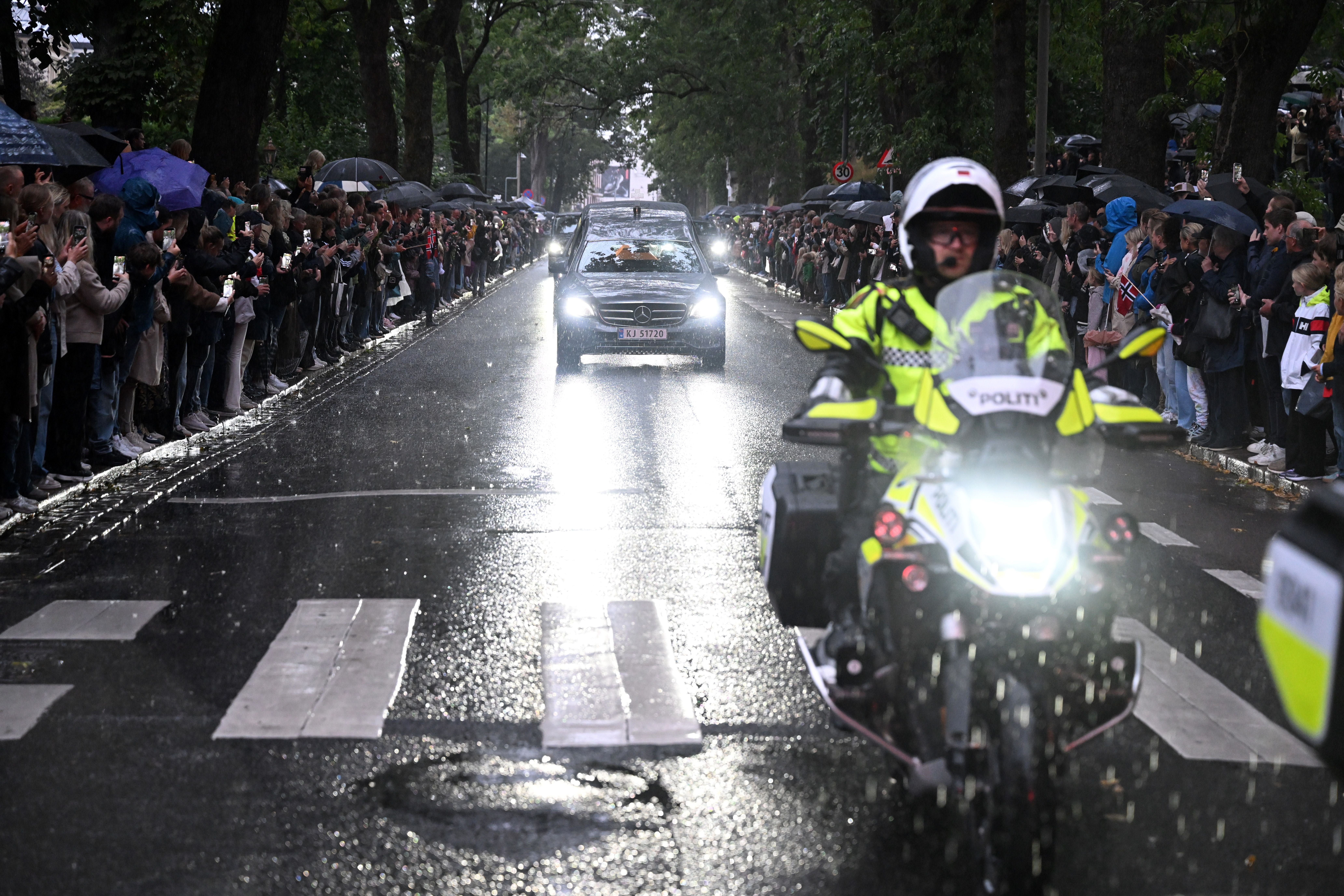 Menschen stehen bei Regen an einer Straße, ein Leichenwagen wird von Polizei und Trauernden begleitet.