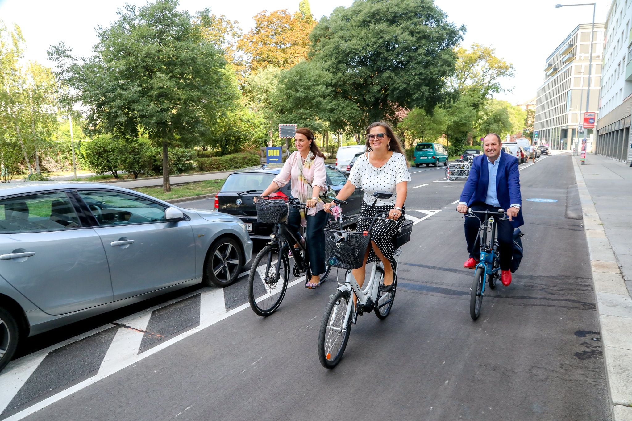 Drei Erwachsene fahren lachend auf Fahrrädern einen Radweg entlang einer Straße in der Stadt.