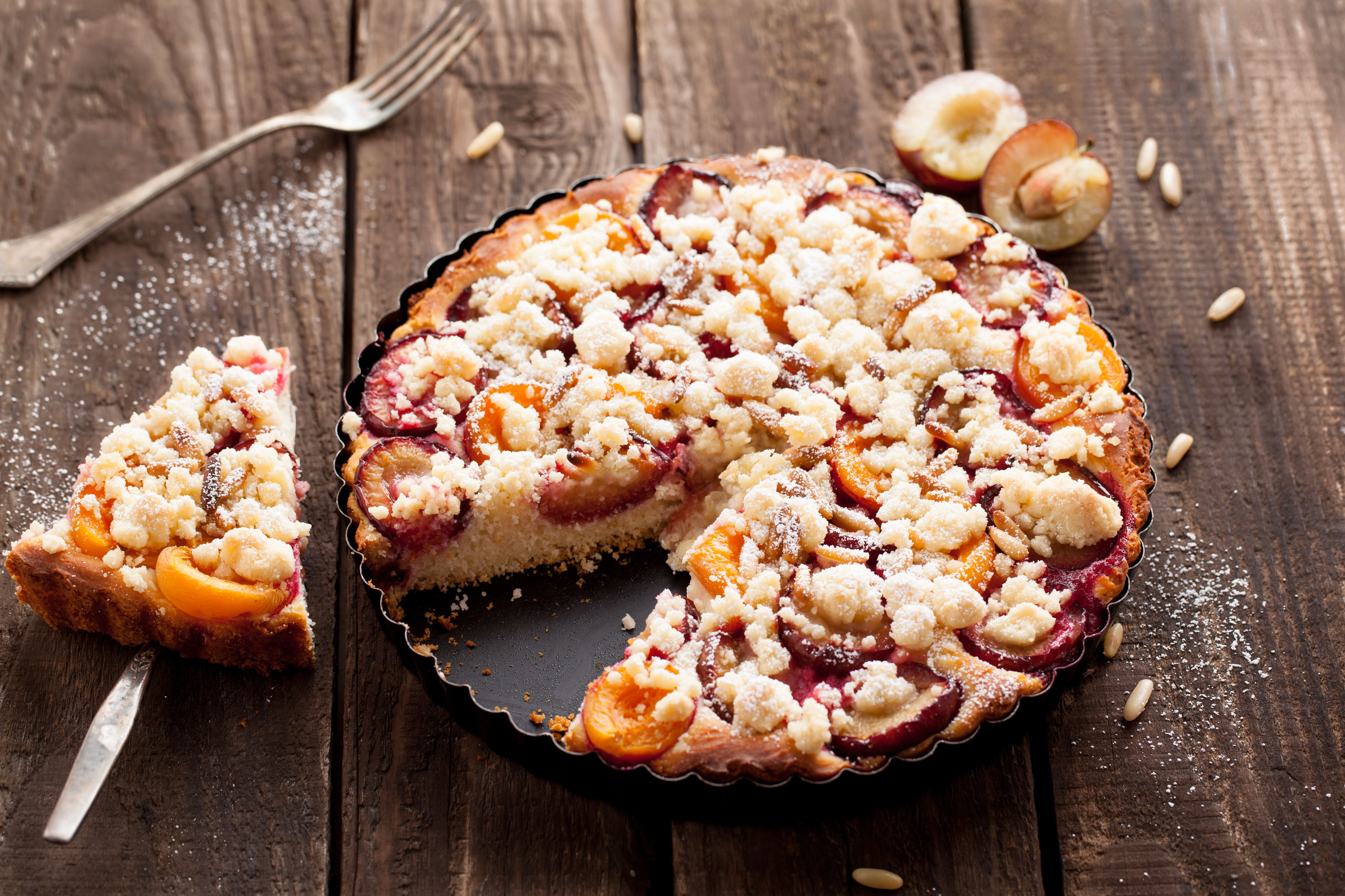 Pflaumenkuchen mit Streuseln und Pinienkernen auf einem Holztisch, ein Stück ist herausgeschnitten.
