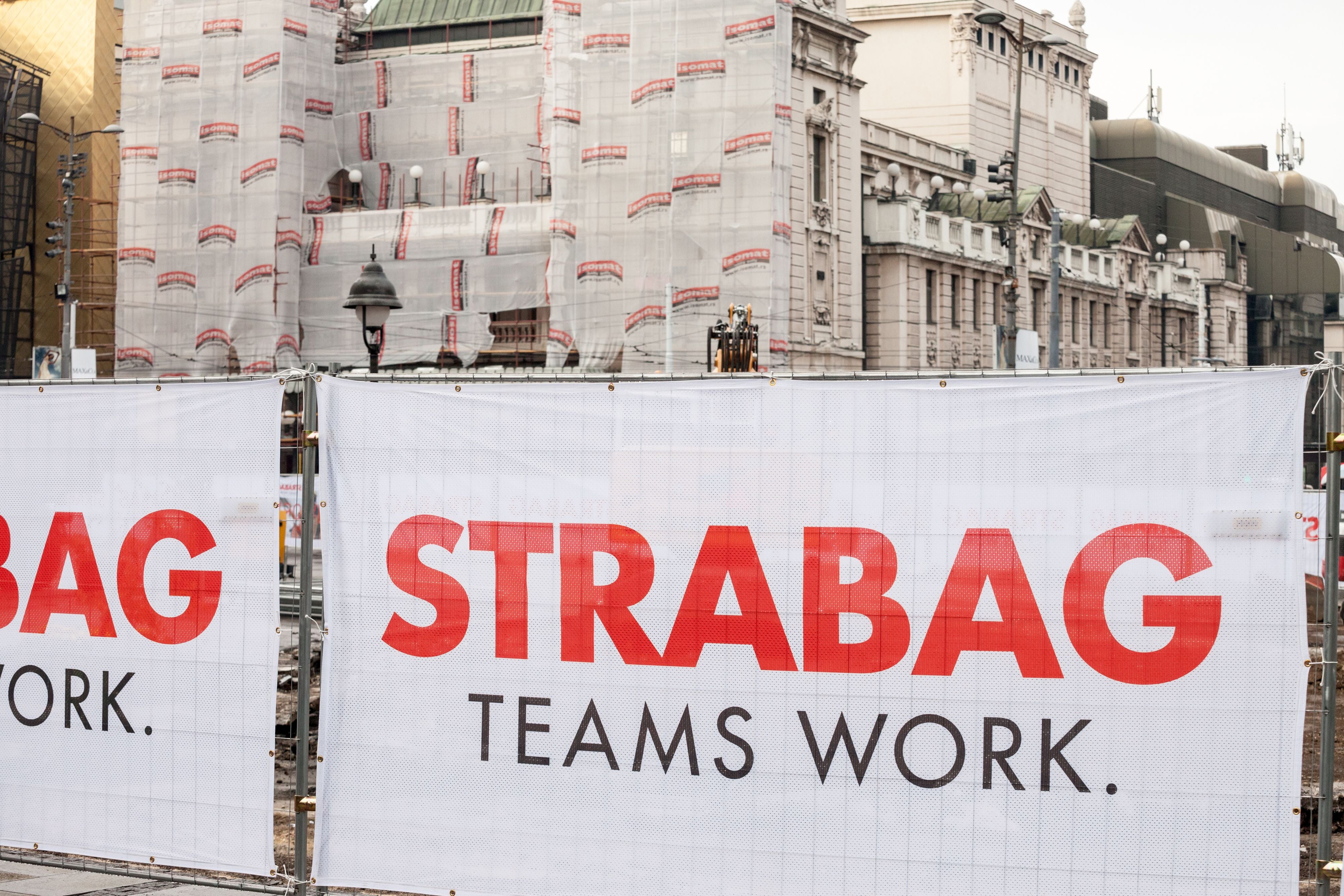 Baustellenabsperrung mit großem STRABAG-Logo und dem Slogan „TEAMS WORK.“ auf einem Bauzaun in einer Straße in Belgrad; im Hintergrund sind Gebäude und Baugerüste zu sehen.