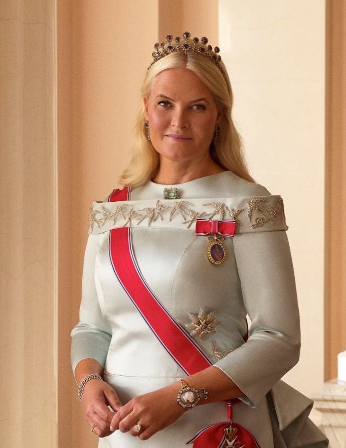 Mette-Marit mit Tiara, elegantem Kleid und Schärpe posiert vor einem hellen Hintergrund.