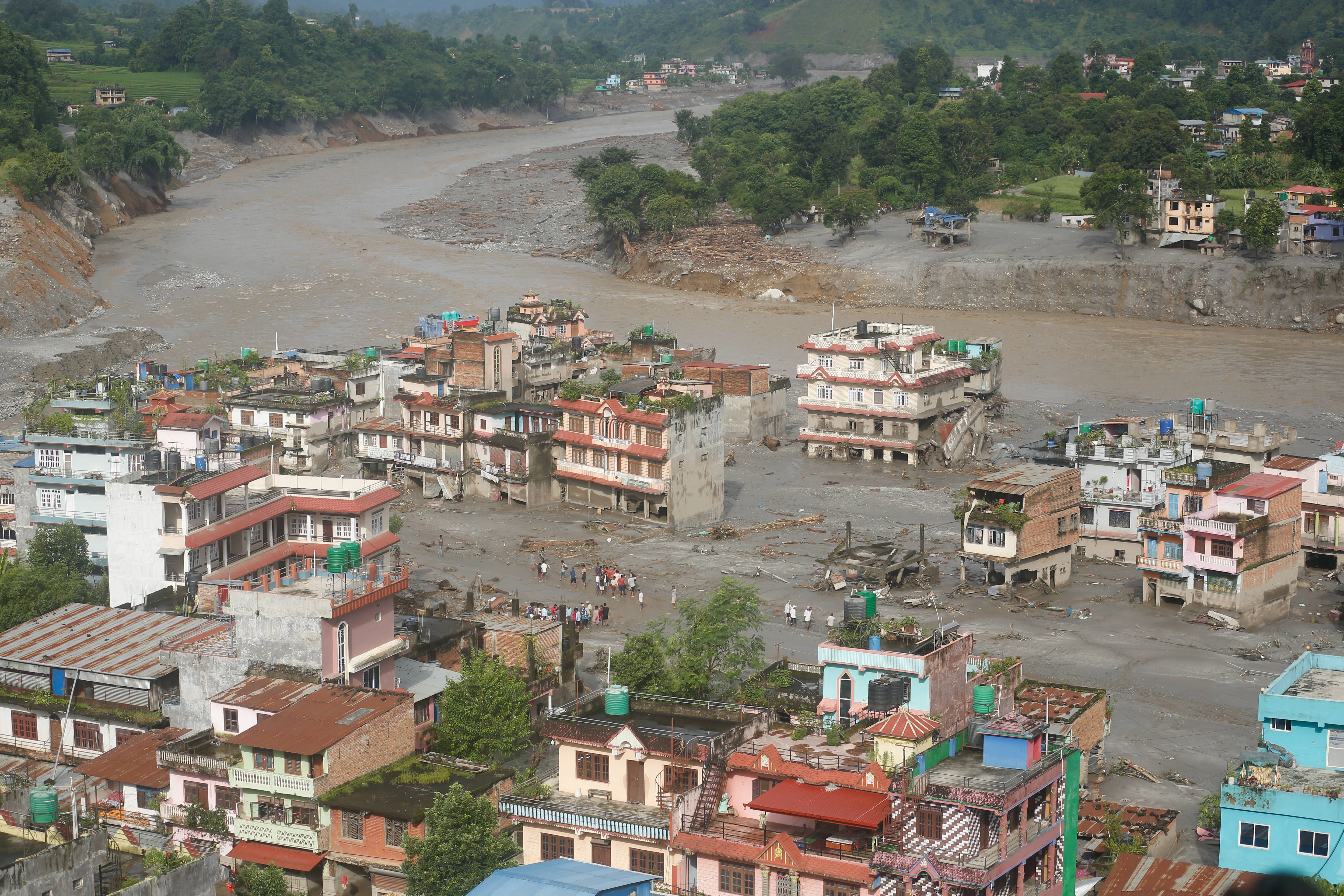 Überflutete Stadt in Nepal mit beschädigten Gebäuden und Menschen auf schlammigen Straßen am Fluss.