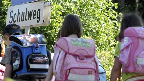 Mehrere Kinder mit Schulranzen gehen unter einem Straßenschild mit der Aufschrift „Schulweg“.