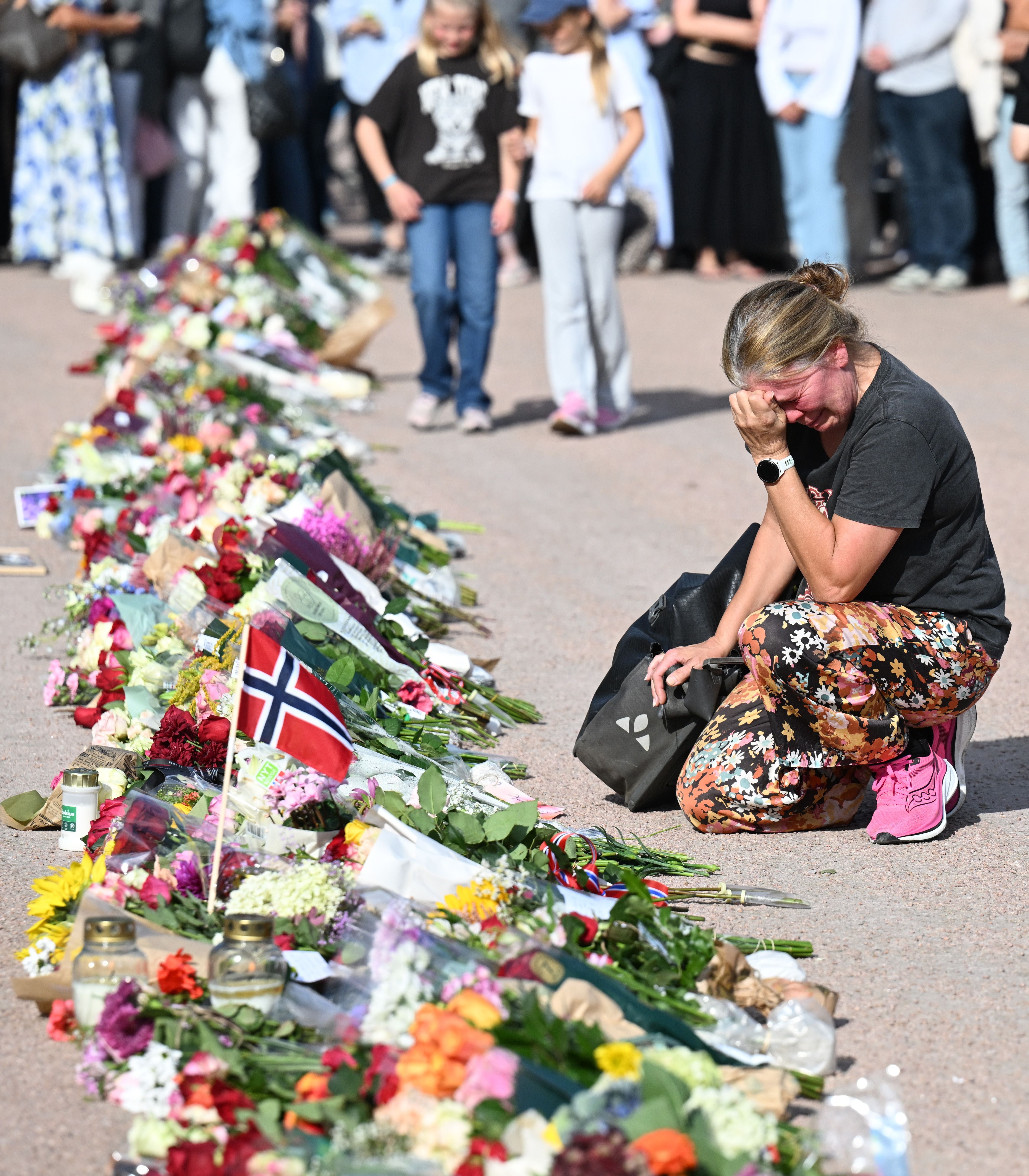 Eine Frau kniet weinend an einem Blumenmeer mit norwegischer Flagge, Menschen im Hintergrund trauern.