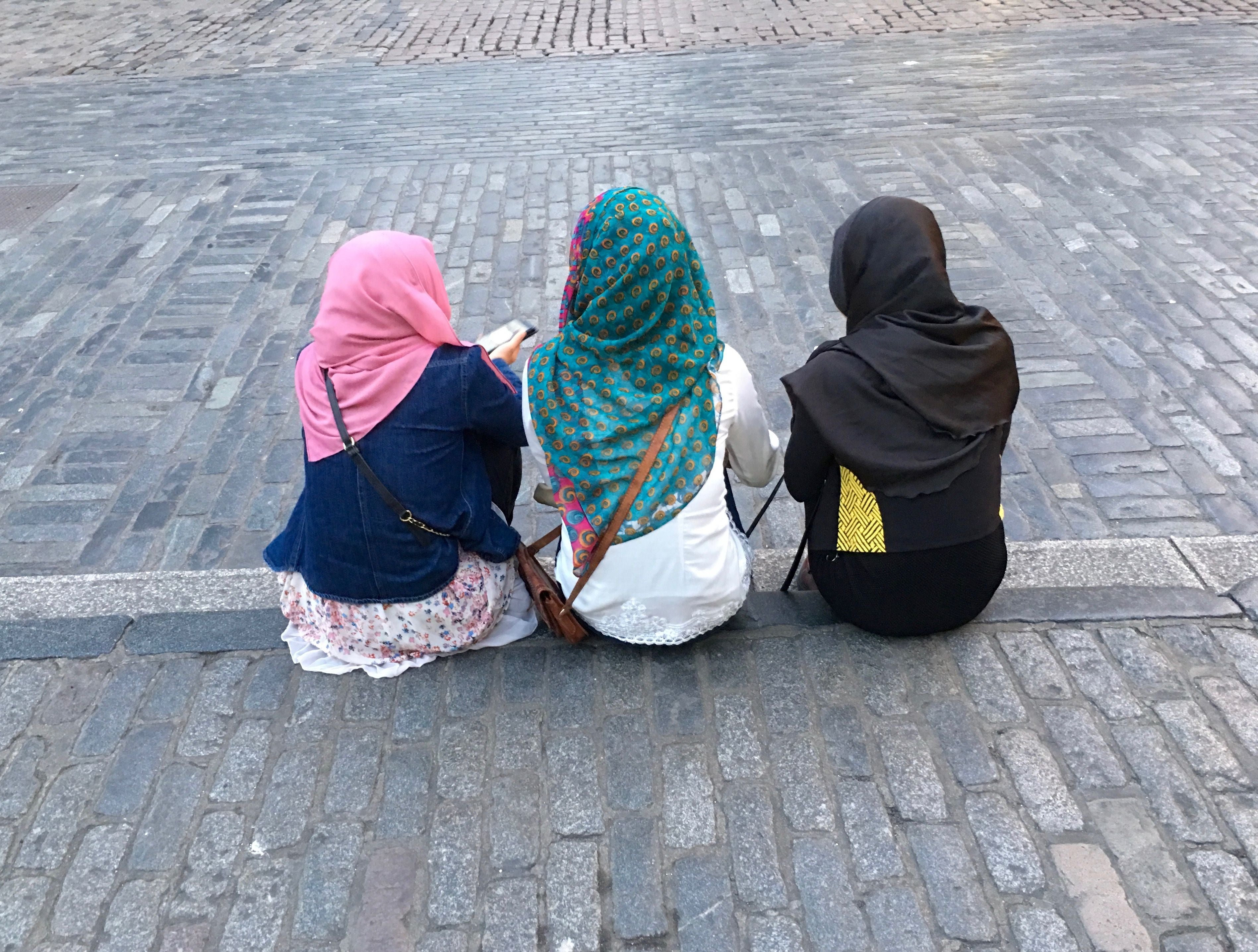 Drei junge Frauen mit Kopftüchern sitzen auf einem Bordstein und nutzen ihre Smartphones.
