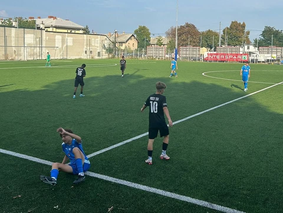 Jugendliche spielen Fußball auf einem Kunstrasenplatz, ein Spieler in Blau sitzt am Boden.