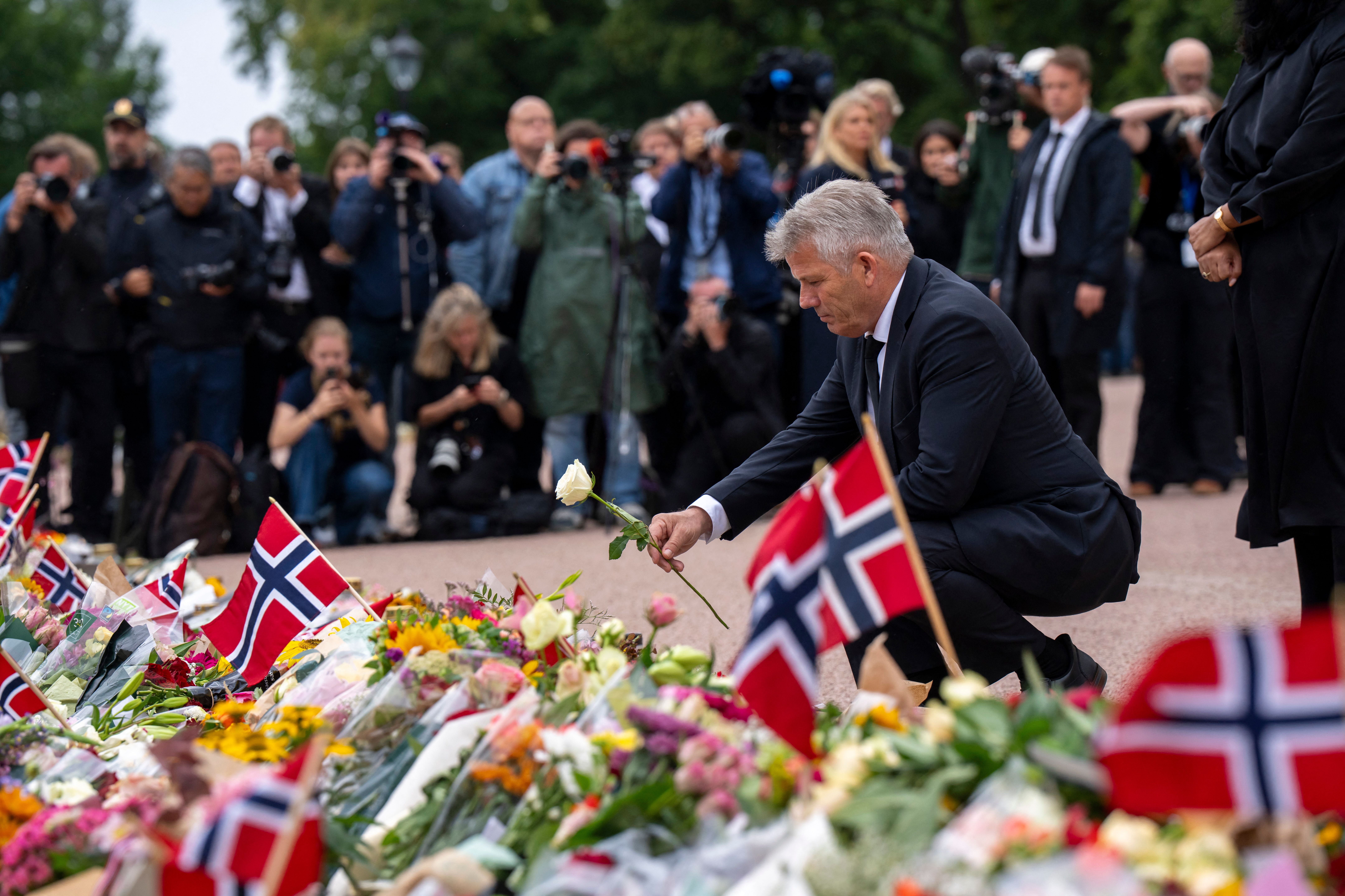 Ein Mann legt eine weiße Rose an ein Blumenmeer mit norwegischen Flaggen, Menschen gedenken.