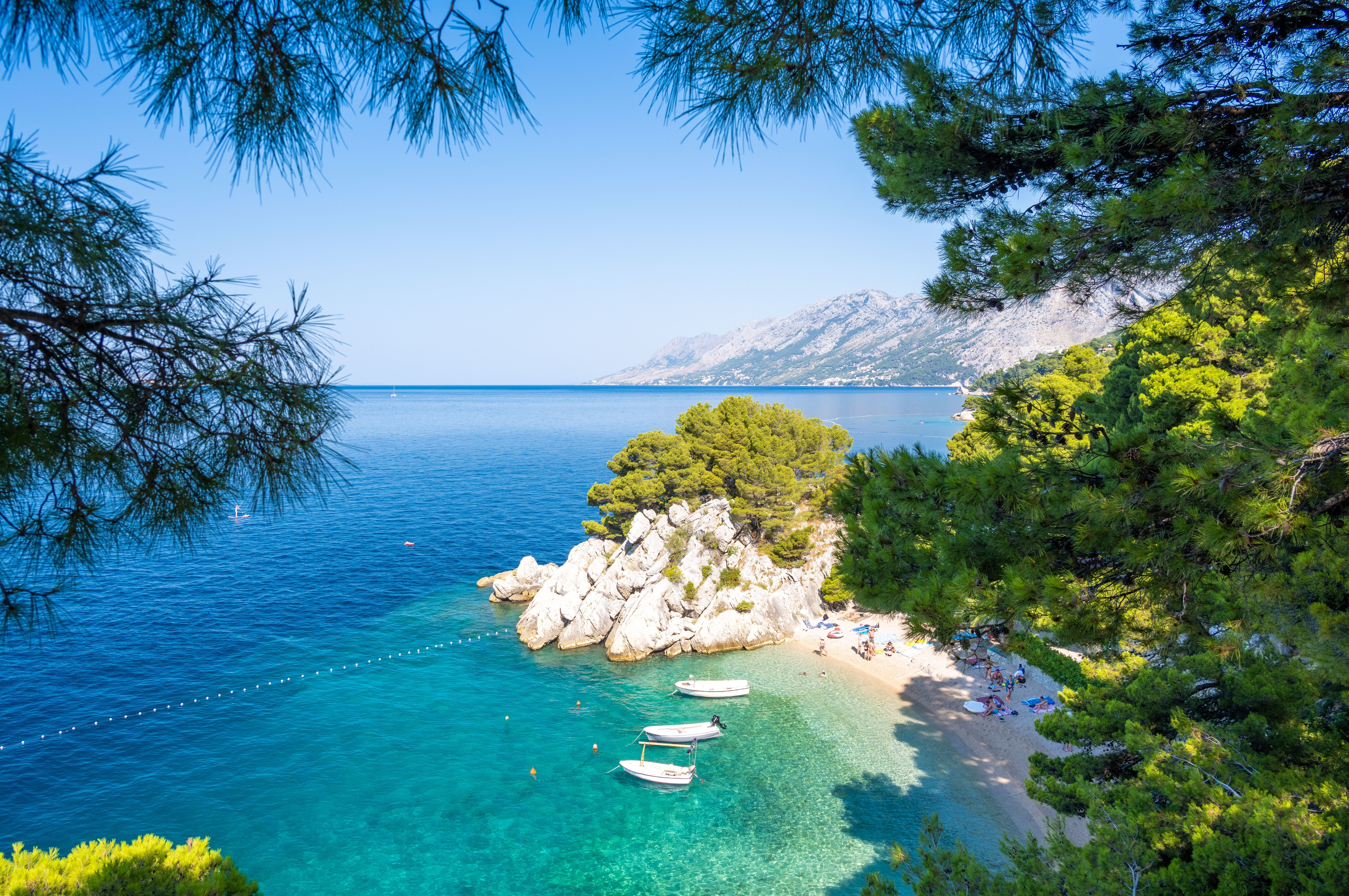 Klares, türkisfarbenes Meer, Sandstrand, Boote und Pinien an der dalmatinischen Küste Kroatiens.