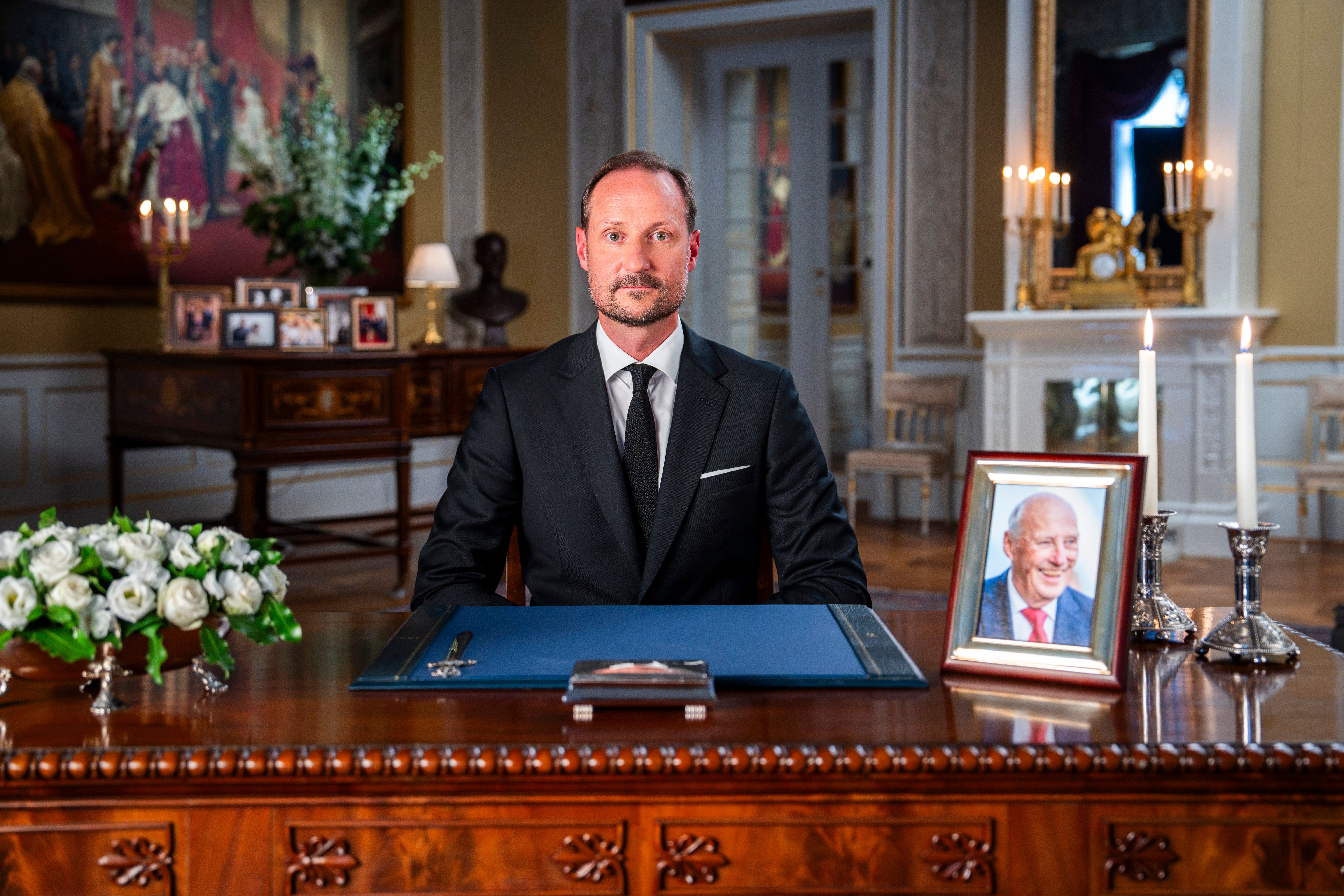 Ein Mann im schwarzen Anzug sitzt an einem Schreibtisch mit Blumen, Kerzen und einem gerahmten Foto.