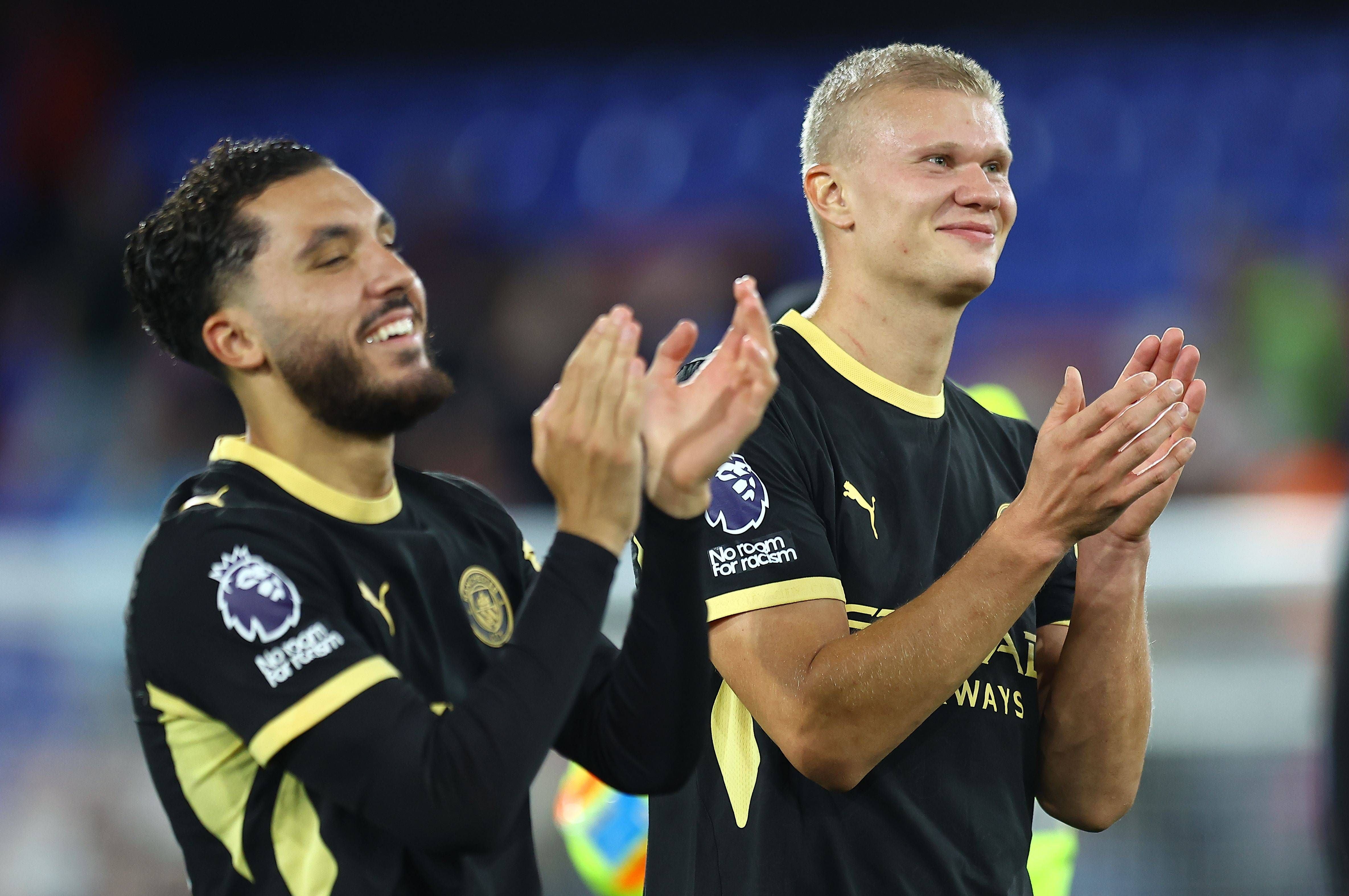 Rayan Cherki und Erling Haaland in schwarzen Trikots mit gelben Akzenten stehen nebeneinander und klatschen lächelnd in die Hände. Der linke Spieler hat dunkles, gelocktes Haar und einen Vollbart, der rechte Spieler hat blonde, kurze Haare.