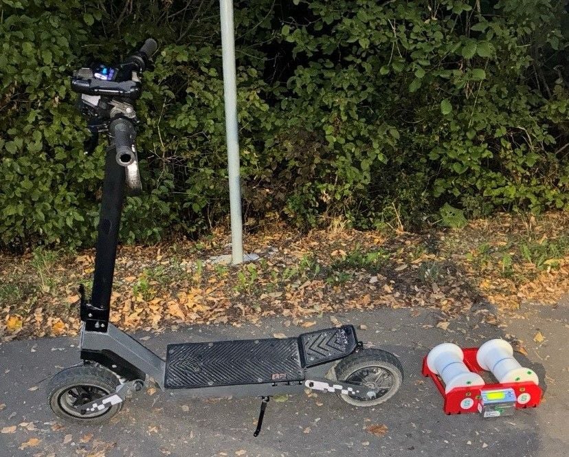 Ein E-Scooter steht auf einem Weg neben einem kleinen roten Messgerät mit weißen Rollen.