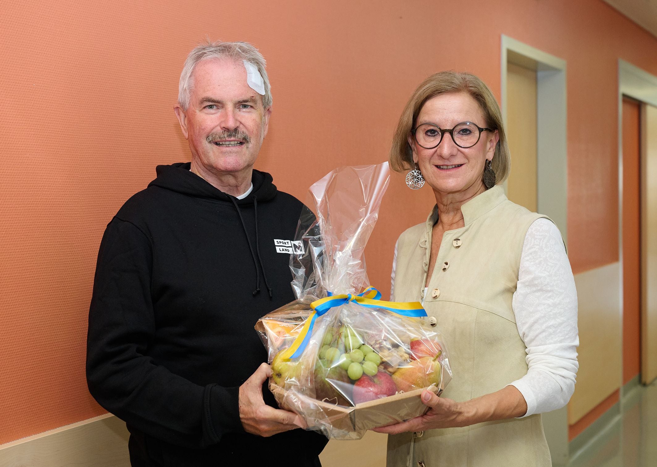 Eine Frau überreicht einem Mann mit Verband ein Obstgeschenkkorb auf einem Flur.