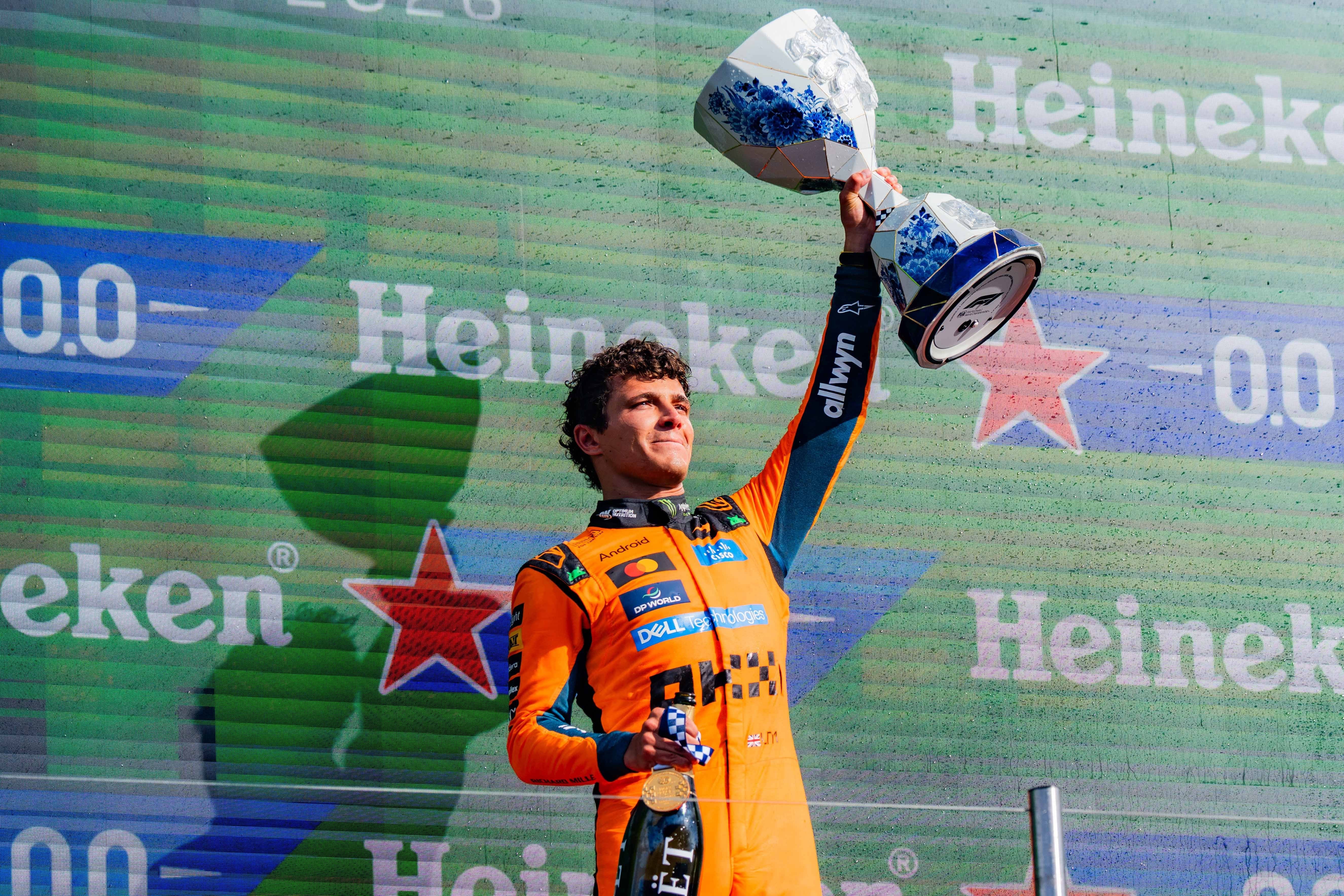 Ein Formel-1-Fahrer im orangen Anzug hält Trophäe und Champagnerflasche jubelnd hoch.