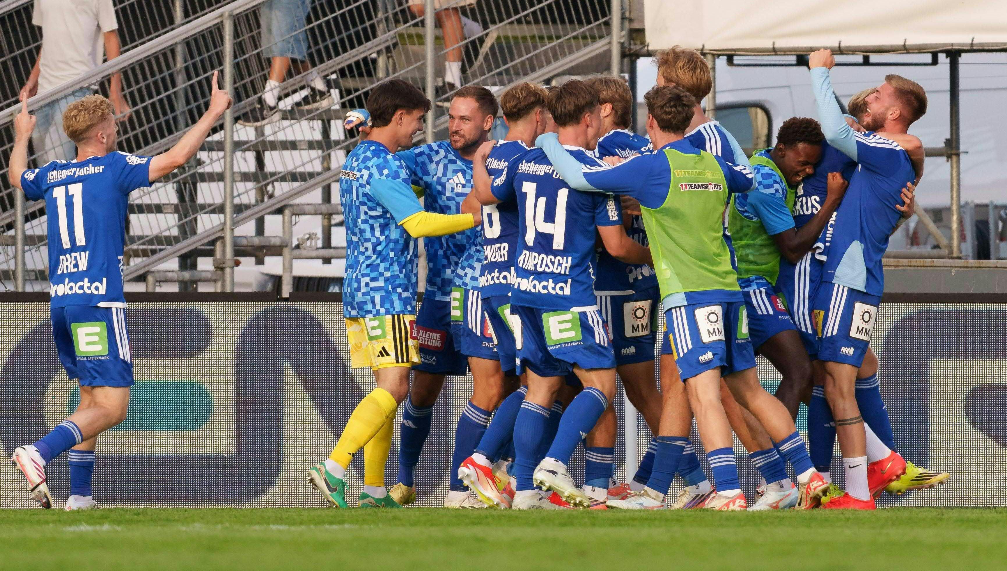 Eine Gruppe von Hartberg-Fußballspielern in blauen Trikots feiert gemeinsam am Spielfeldrand vor einer Werbebande und Tribünenkonstruktion. Die Spieler umarmen sich jubelnd, während ein Spieler mit der Rückennummer 11 mit erhobenem Arm herbeiläuft.