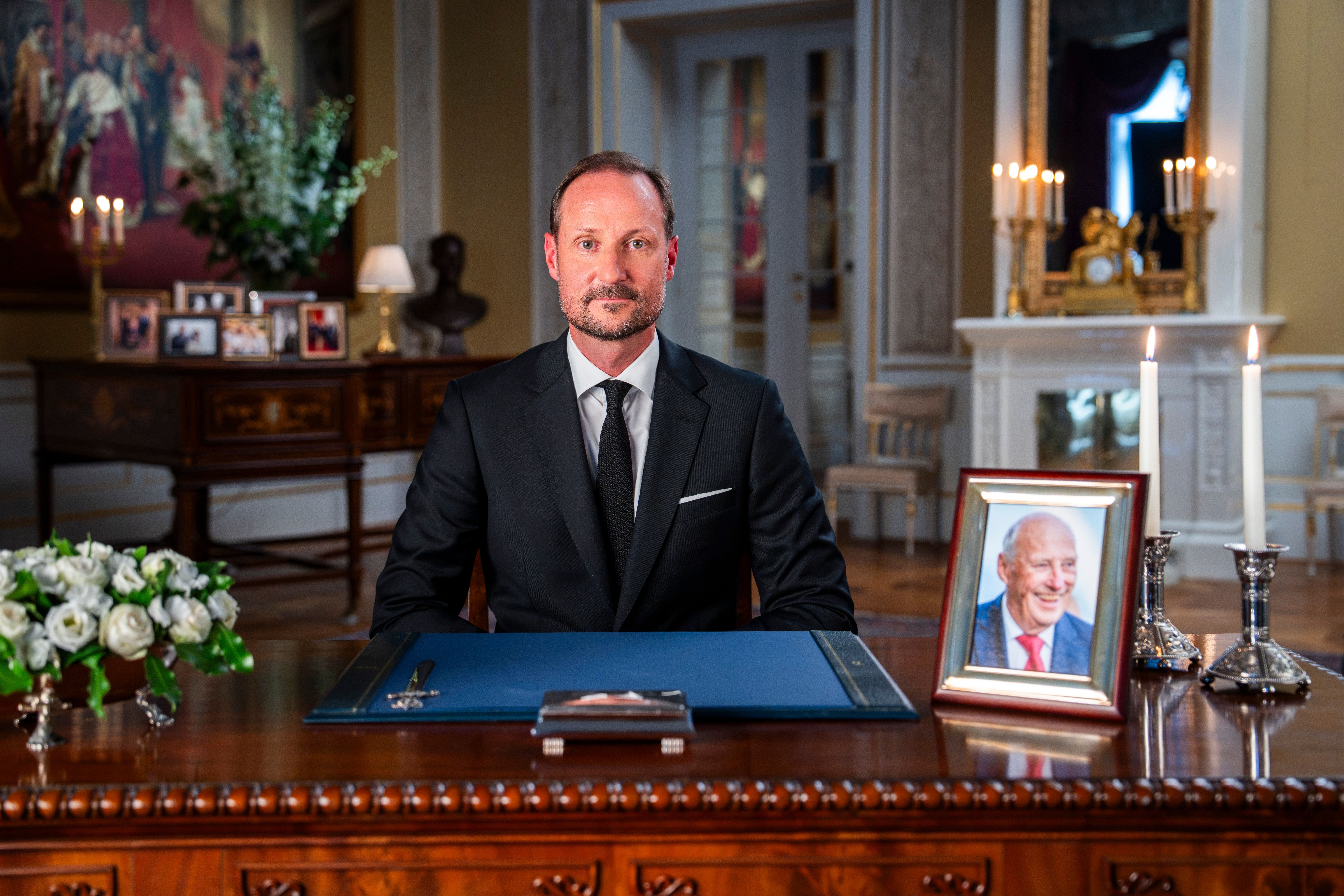 Ein Mann im Anzug sitzt an einem Schreibtisch mit Foto eines älteren Mannes und brennenden Kerzen.