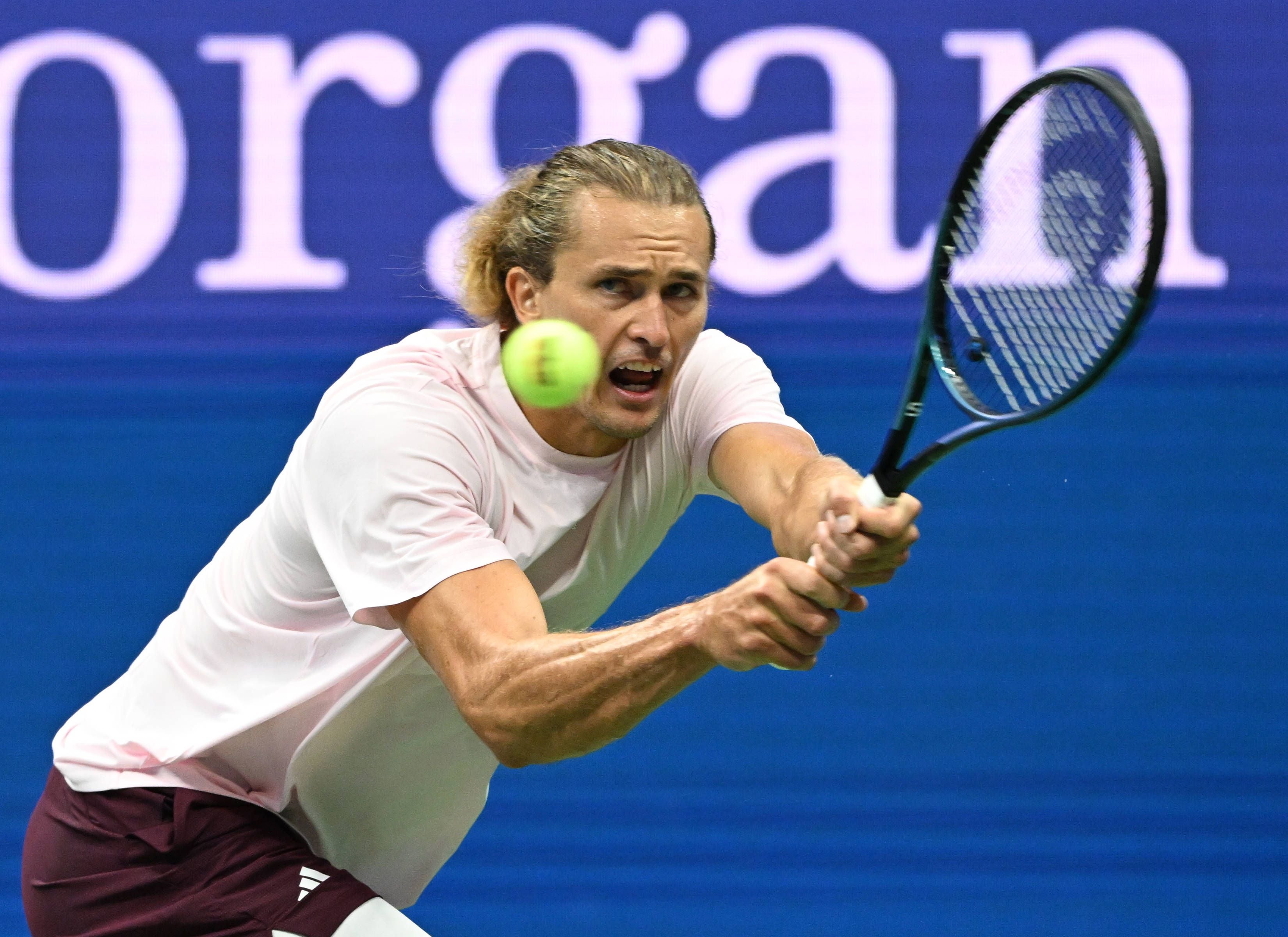 Tennisspieler Alexander Zverev mit längeren blonden Haaren im rosafarbenen T-Shirt schlägt den gelben Tennisball mit beidhändiger Rückhand auf einem blauen Hartplatz.