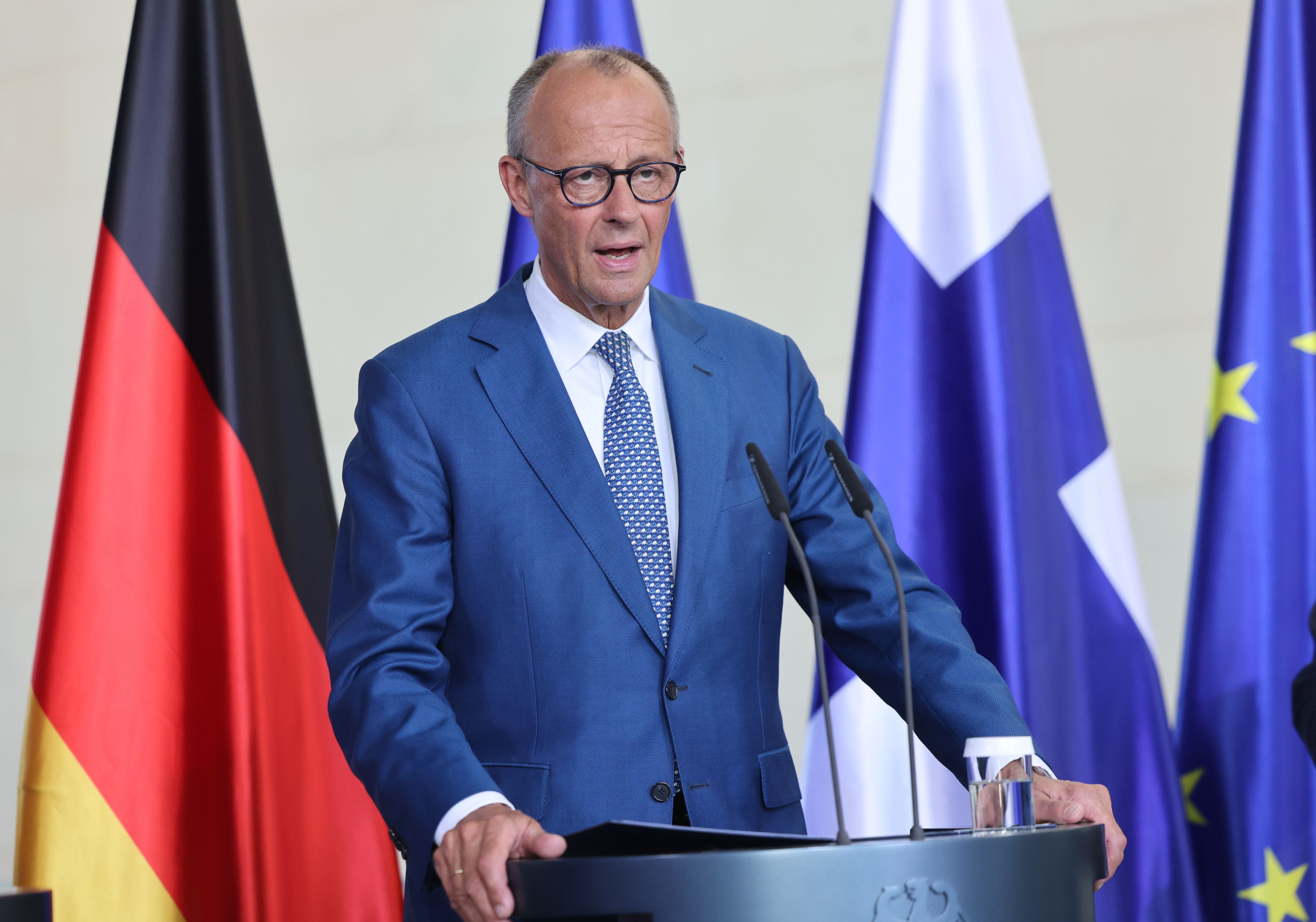 Ein Mann im blauen Anzug spricht auf einer Pressekonferenz vor mehreren Nationalflaggen.