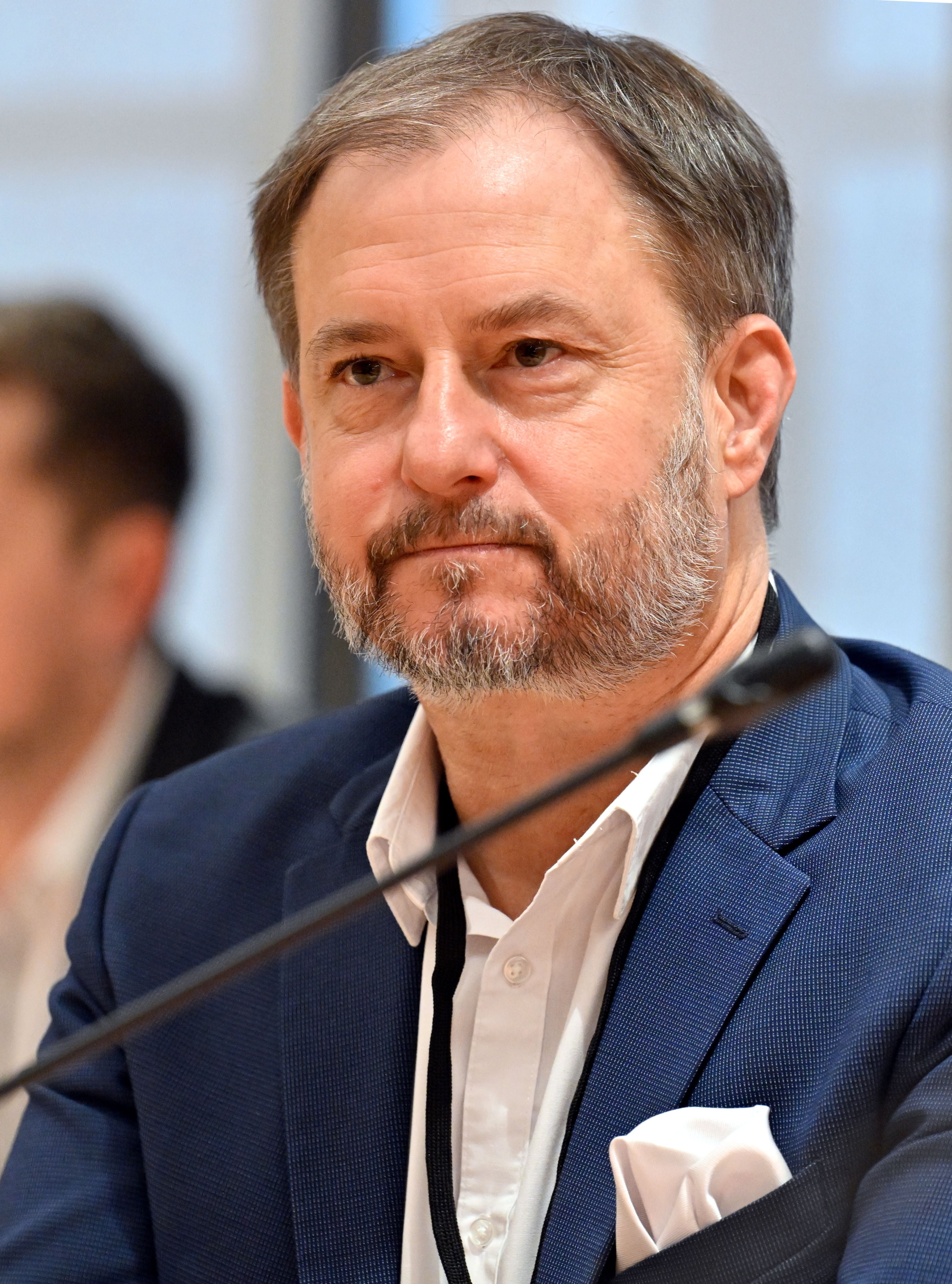 Ein Mann im blauen Sakko sitzt bei einer Veranstaltung hinter einem Mikrofon.