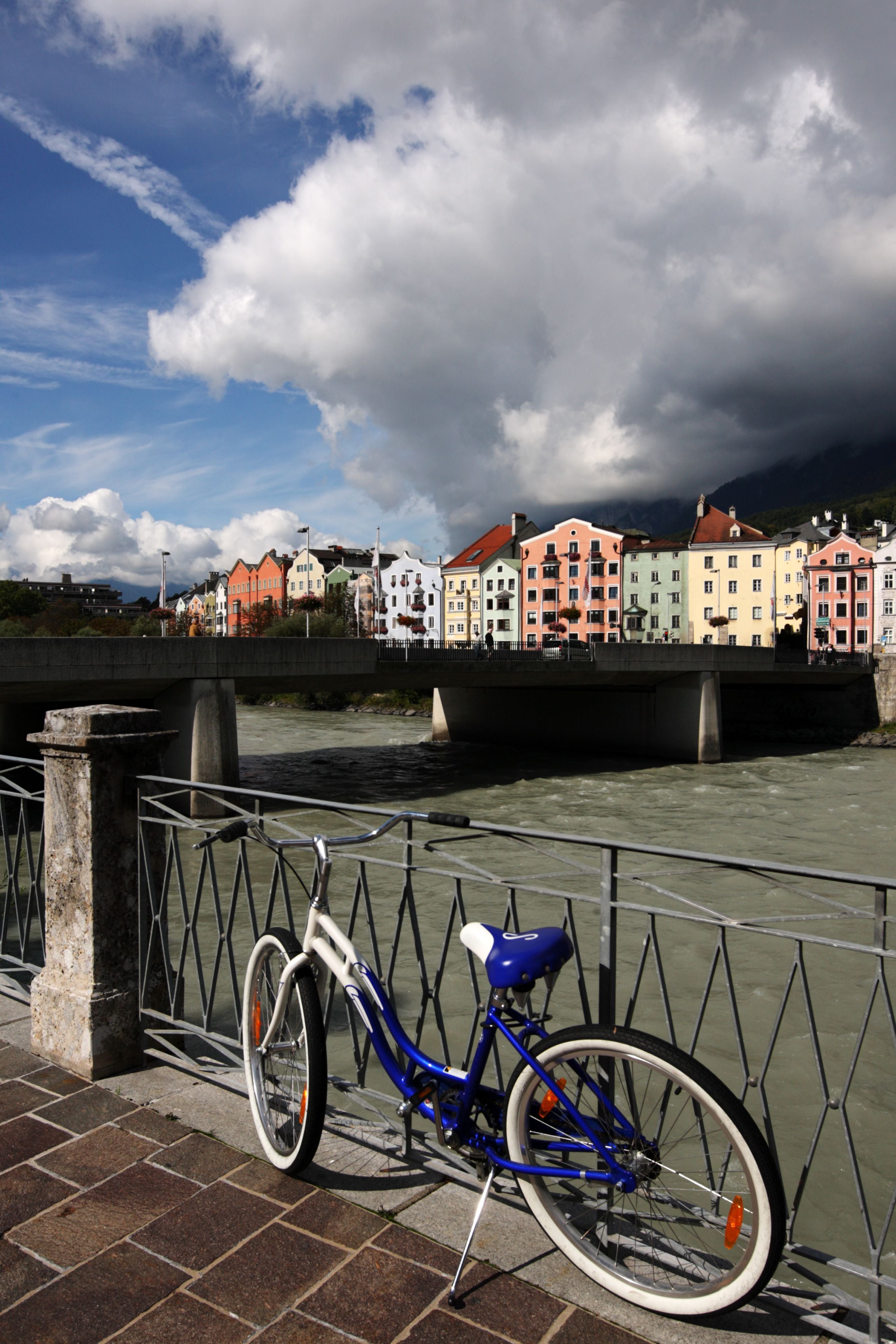Blau-weißes Fahrrad am Inn mit bunten Häusern und dramatischem Himmel in Innsbruck, Österreich.