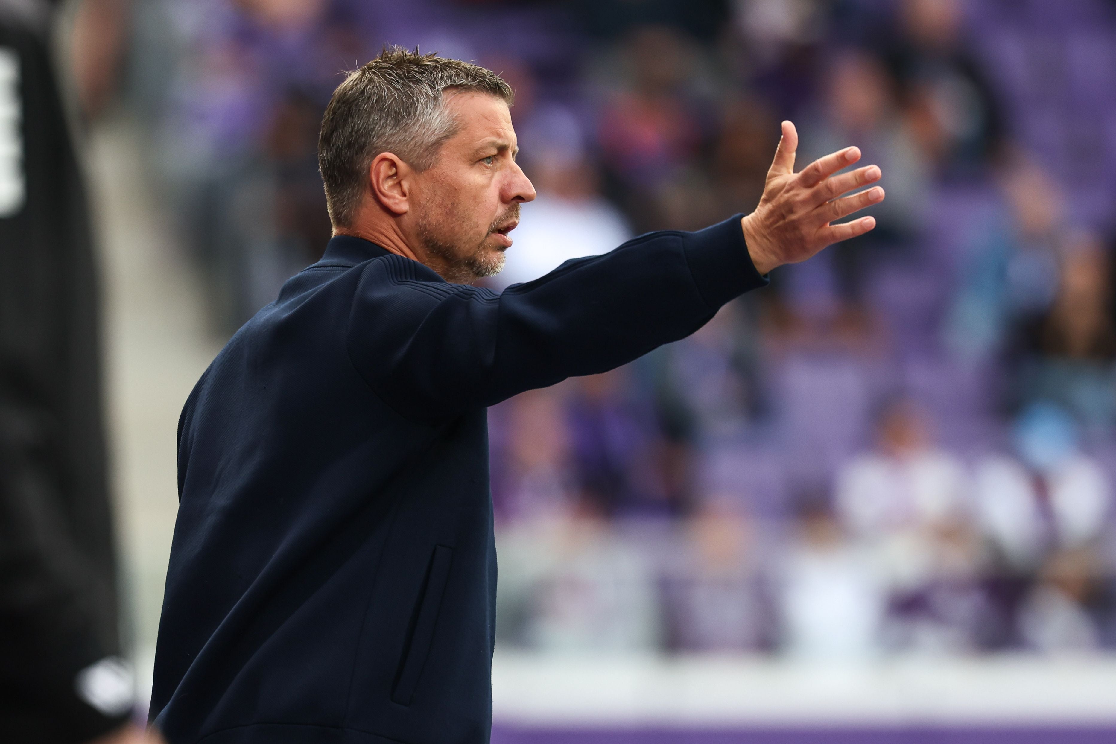 Ein Mann mit kurzen, ergrauenden Haaren und einem dunklen Oberteil steht im Profil und gestikuliert mit ausgestrecktem Arm und geöffneter Handfläche nach rechts. Im Hintergrund ist eine unscharfe Stadionkulisse mit violetten Rängen zu erkennen.