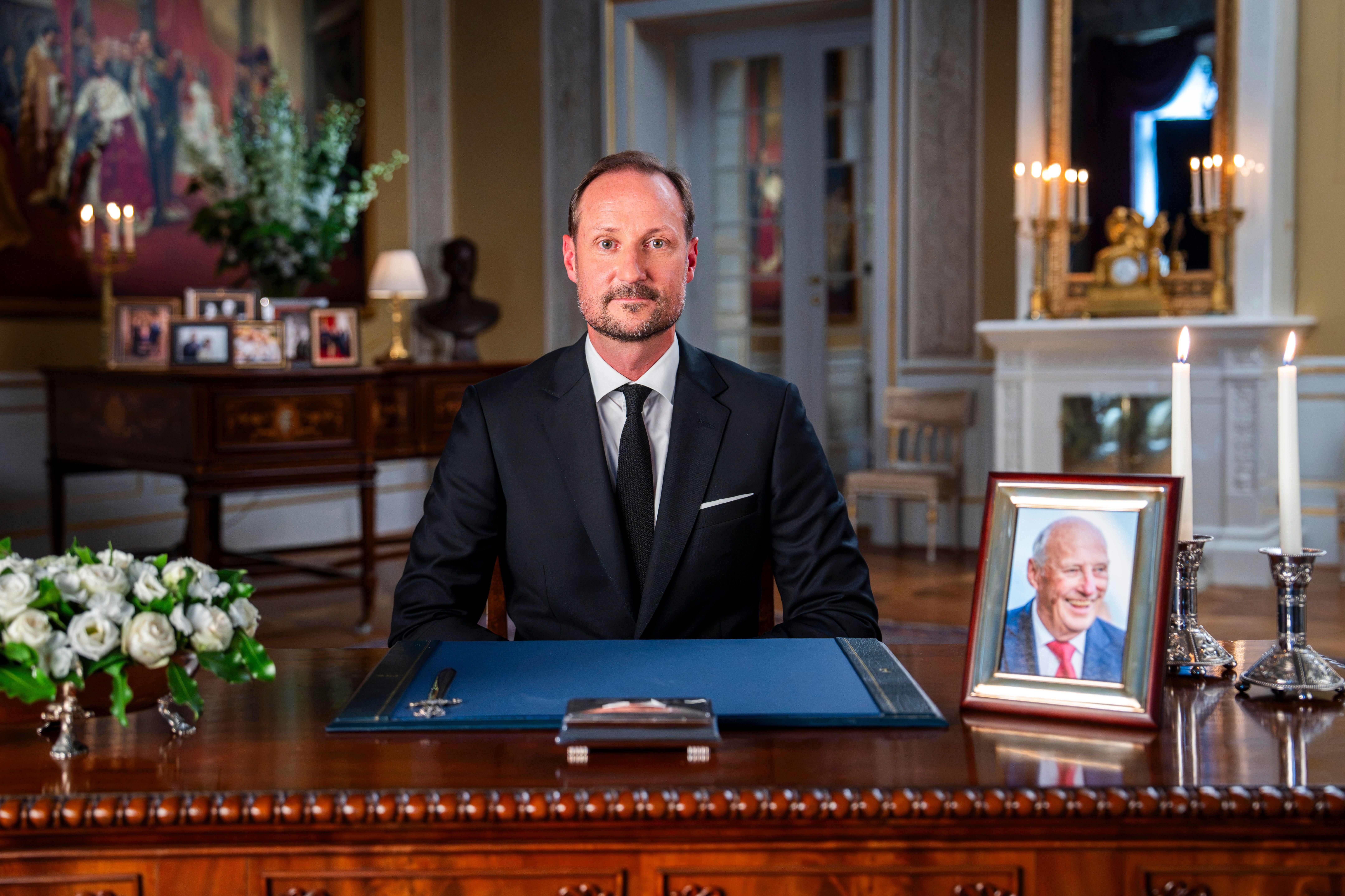 Ein Mann im Anzug sitzt an einem Schreibtisch mit Blumen, Kerzen und gerahmtem Foto eines älteren Mannes.