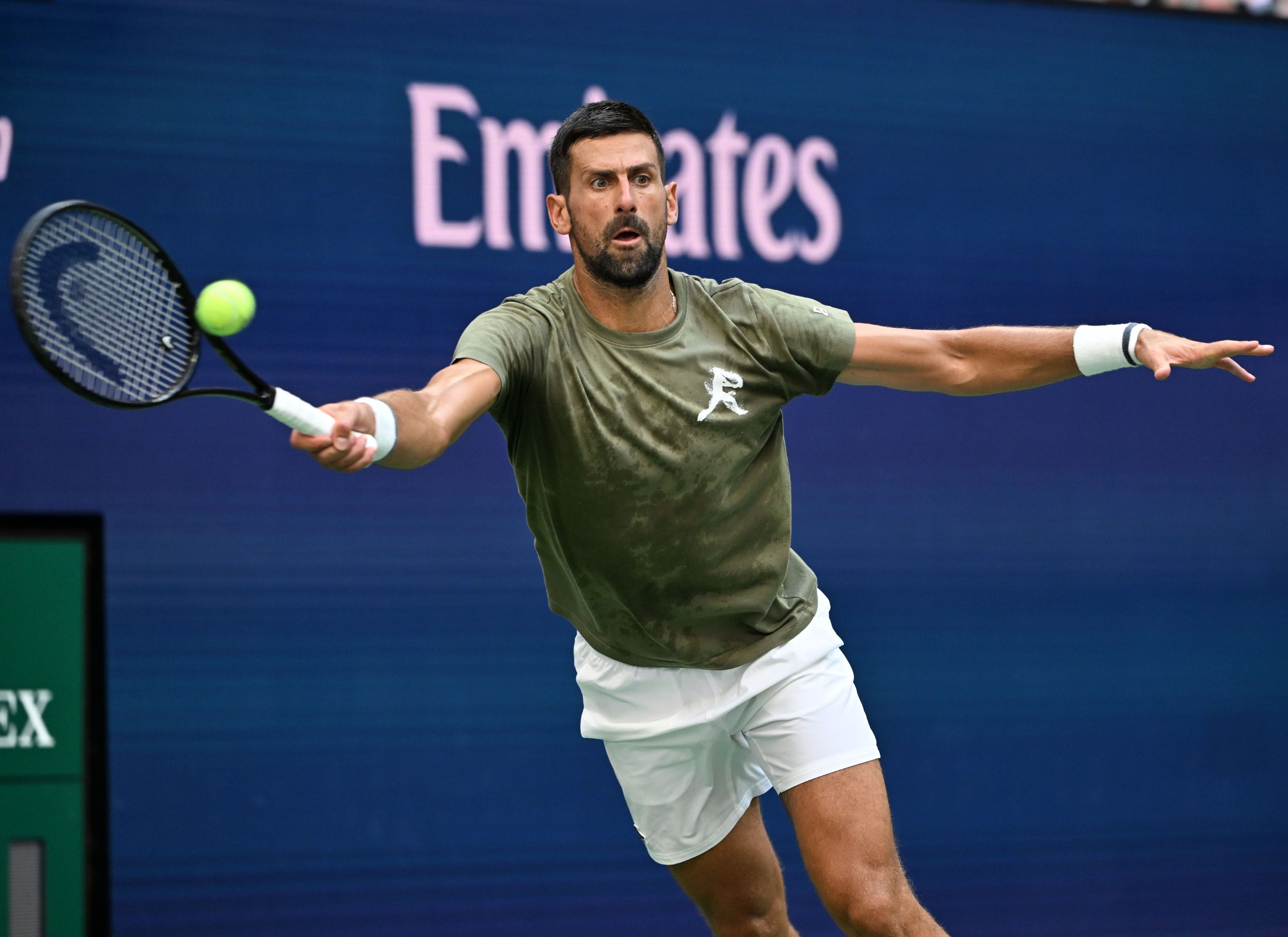 Tennisspieler&nbsp;Novak Djokovic mit Bart in einem olivgrünen T-Shirt und weißer Hose schlägt mit einer Vorhand den Filzball auf einem Hartplatz.