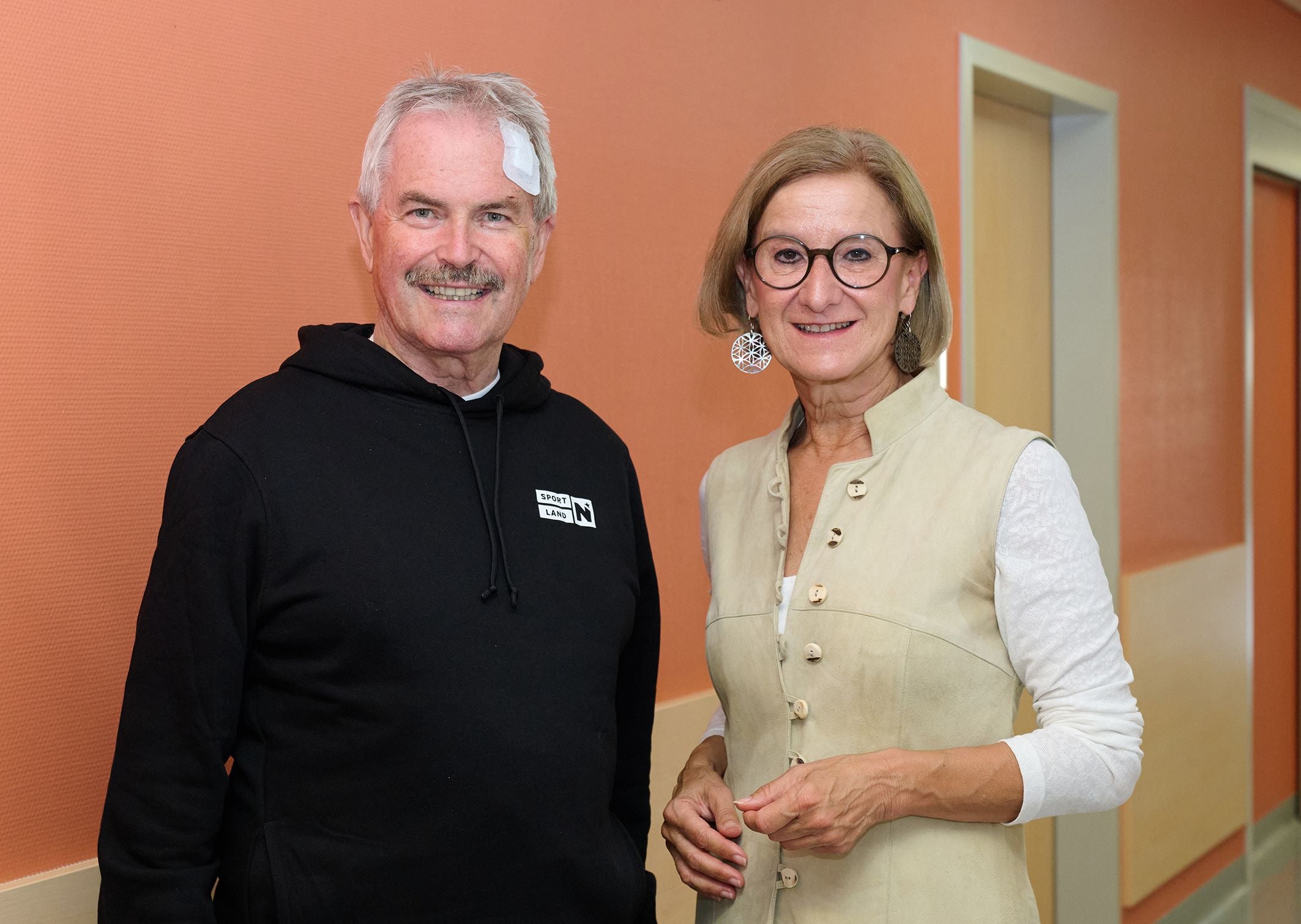 Eine Frau überbringt einem Mann mit Stirnverband Genesungswünsche in einem Flur.
