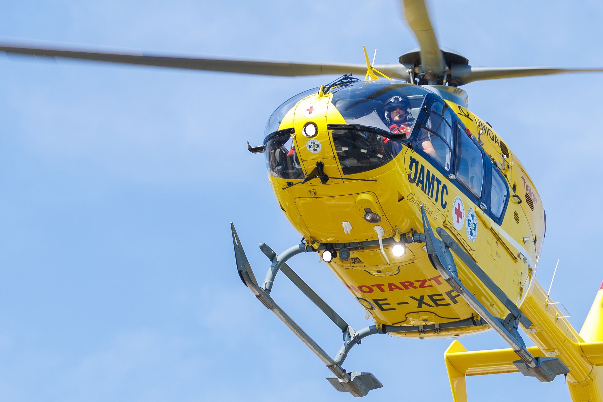 Gelber Rettungshubschrauber des ÖAMTC im Flug vor blauem Himmel, Pilot sichtbar.