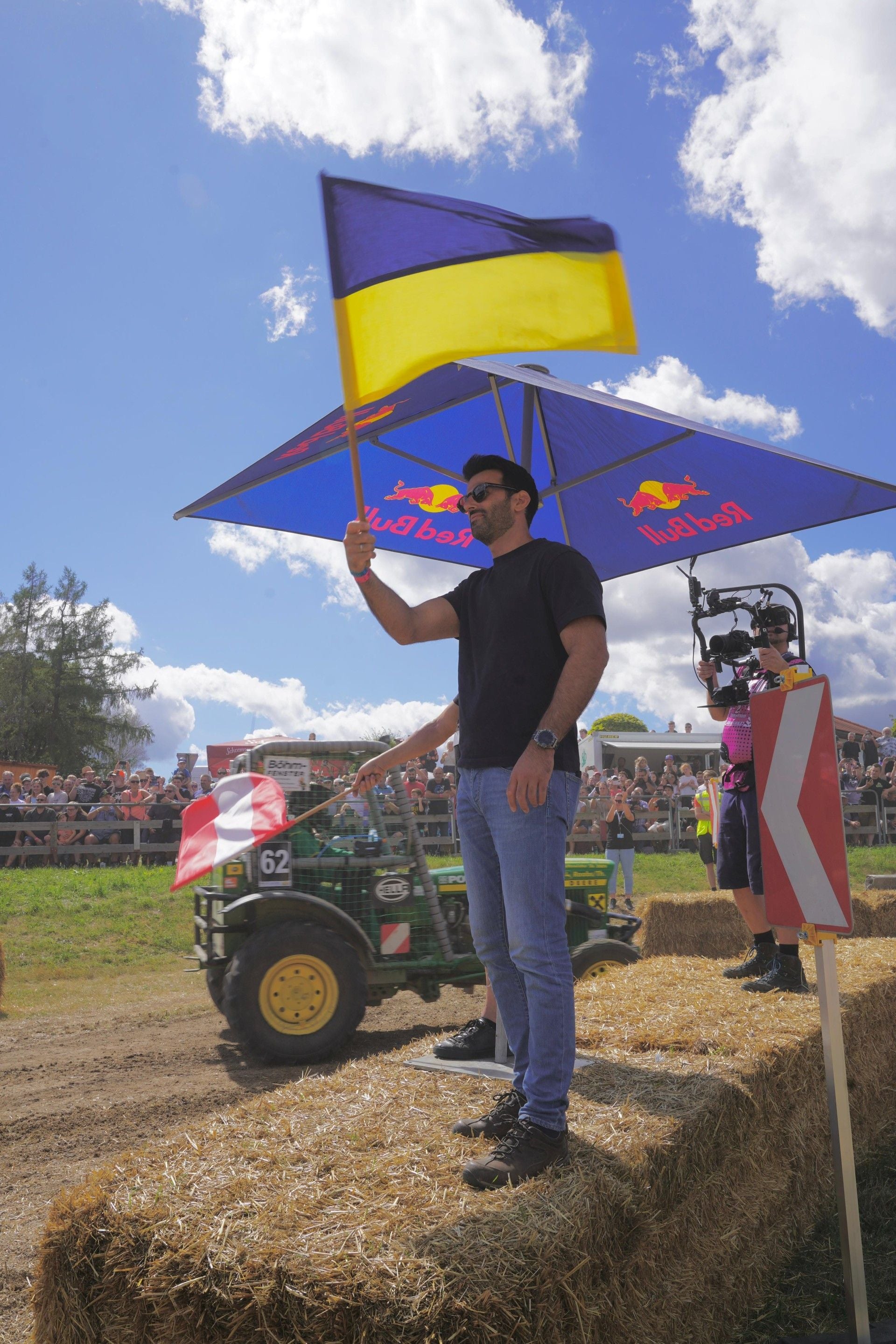 Ein Mann schwenkt eine Flagge beim Traktorenrennen, Zuschauer und Kameramann im Hintergrund.