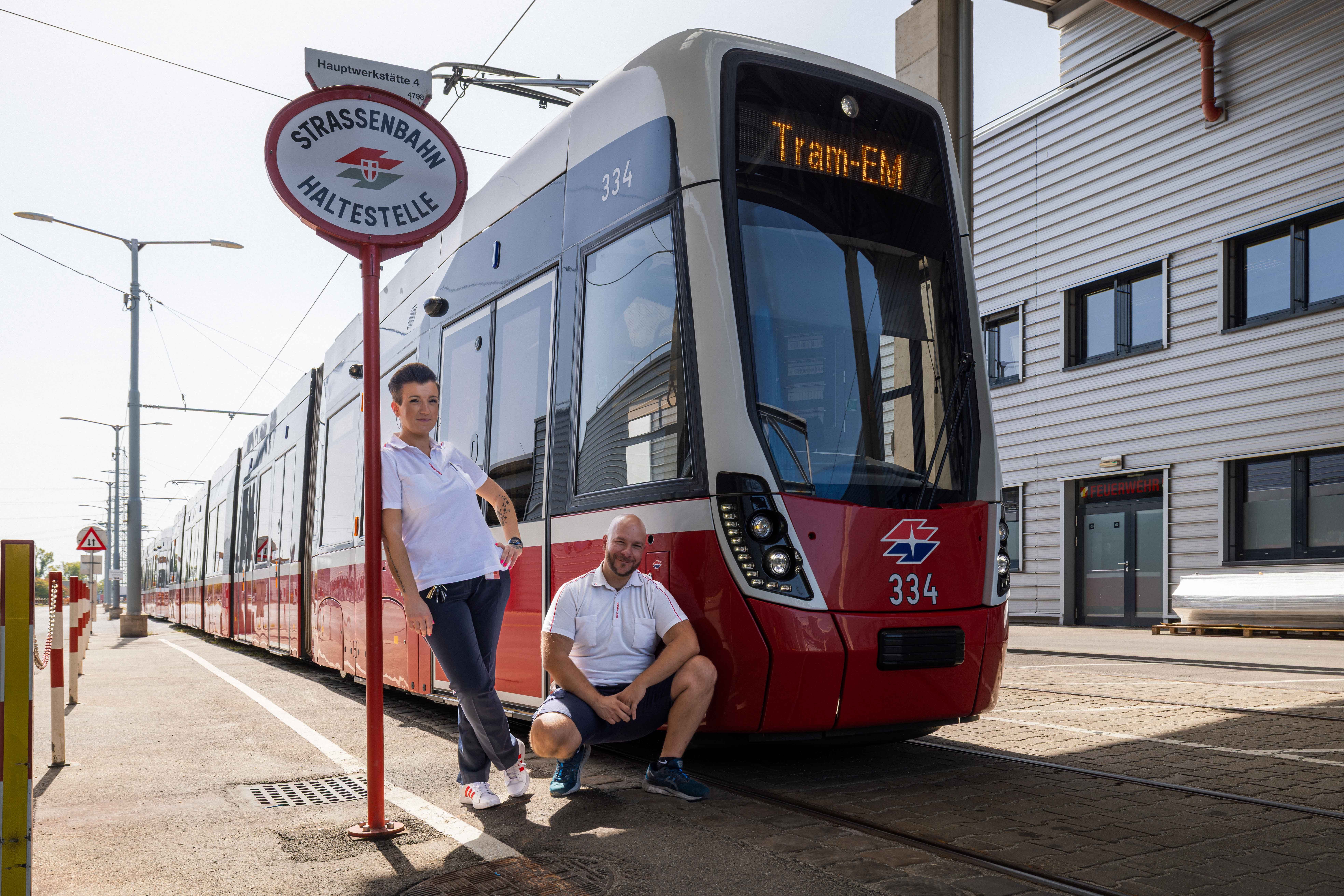 Zwei Personen posieren vor einer Straßenbahn an einer Haltestelle mit dem Schild "STRASSENBAHN HALTESTELLE".