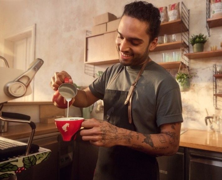 Ein Barista gießt Milch in eine rote Tasse und lächelt in einem gemütlichen Café.