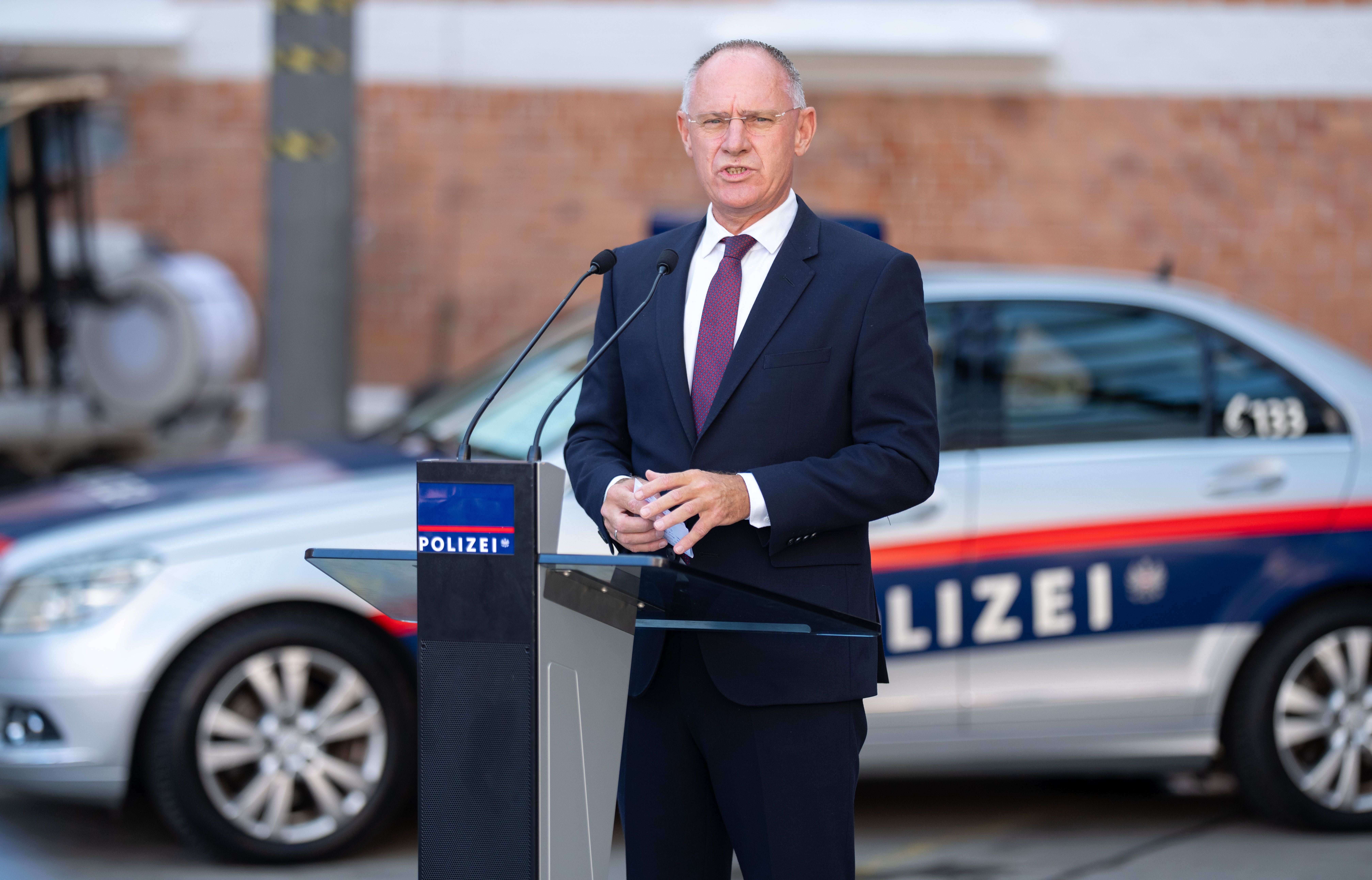 Ein Mann im Anzug spricht an einem Rednerpult vor einem Polizeiauto.