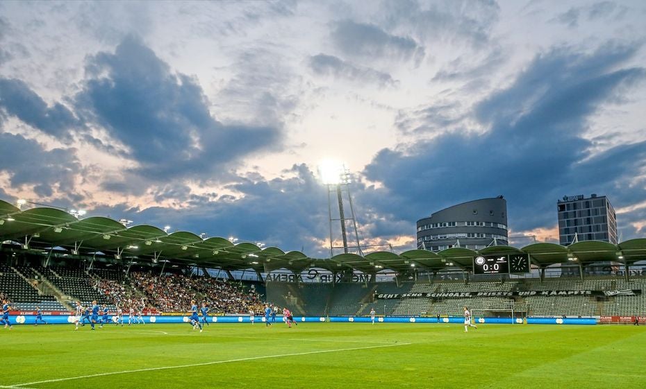 Fußballspiel im Stadion bei Sonnenuntergang, mit Zuschauern auf den Rängen und modernen Gebäuden im Hintergrund.