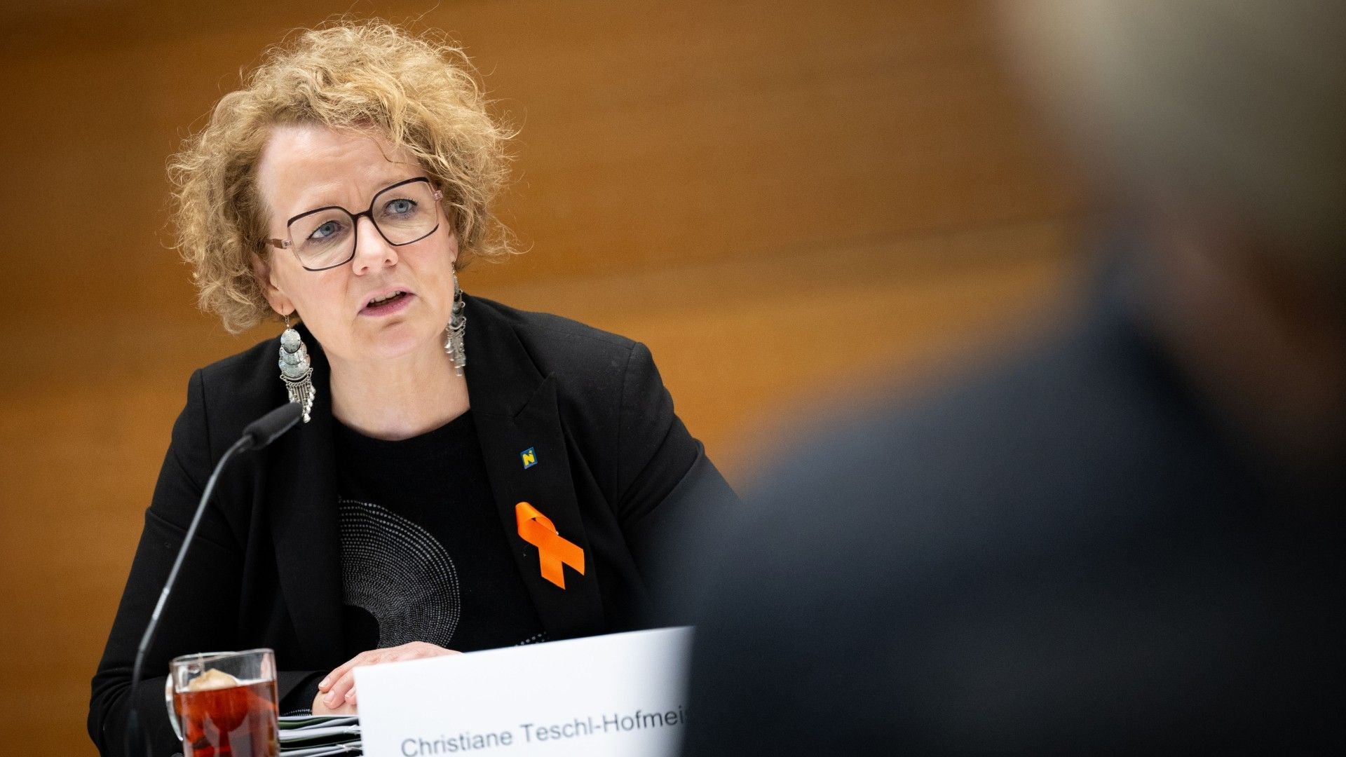 Eine Frau mit Brille und Locken spricht bei einer Veranstaltung, orange Schleife am Sakko.