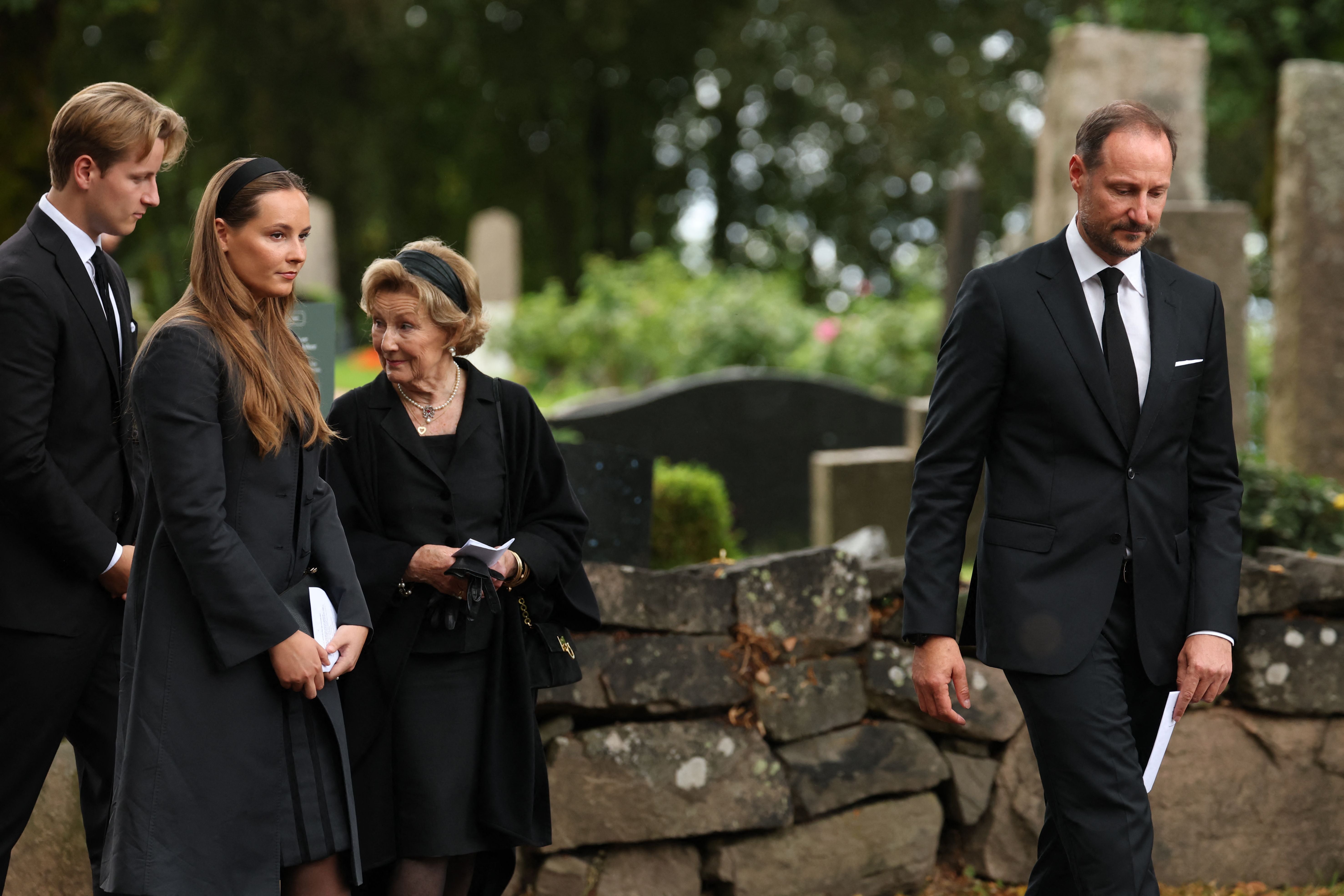 Vier Menschen in schwarzer Trauerkleidung gehen an einem Friedhof entlang.