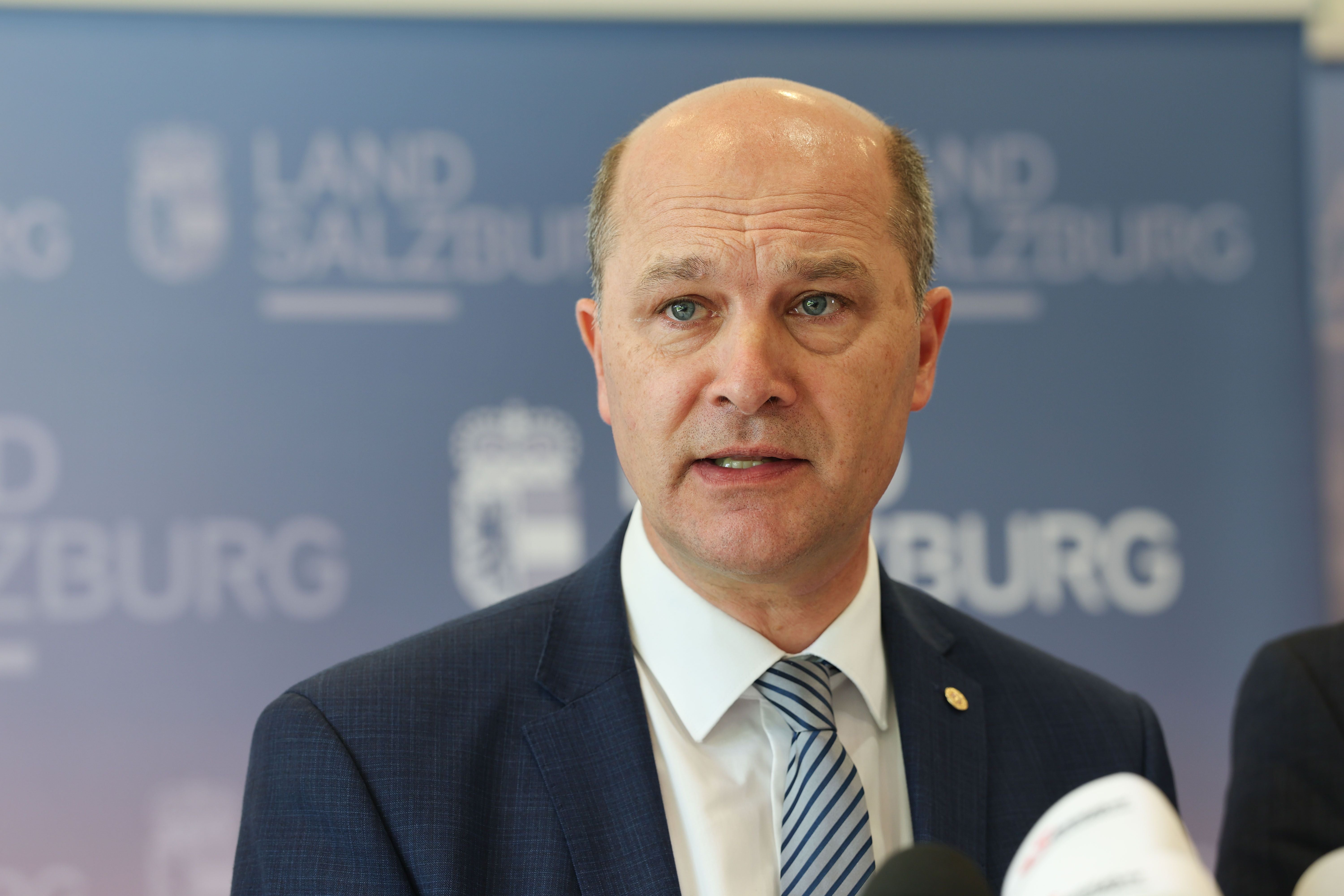 Ein Mann im Anzug spricht bei einer Pressekonferenz vor einem Salzburg-Hintergrund.