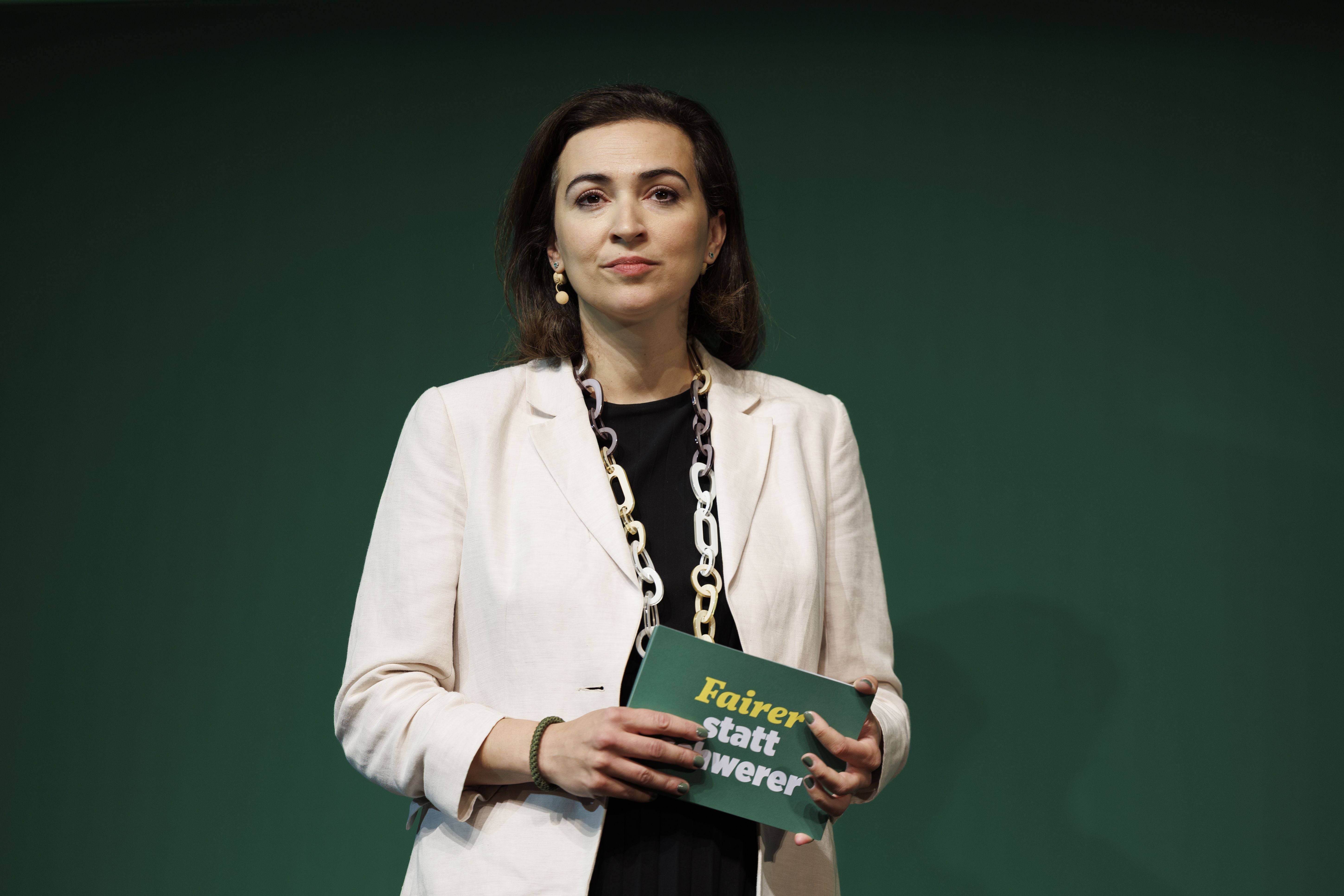 Frau in hellem Blazer hält Schild mit Aufschrift vor grünem Hintergrund.